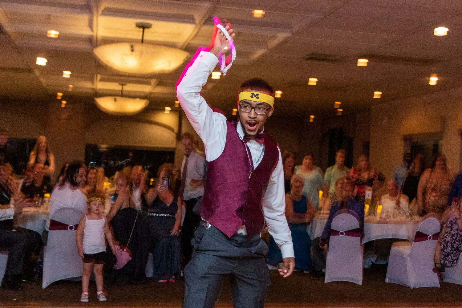 A groom in a burgundy vest dance energetically at a reception.