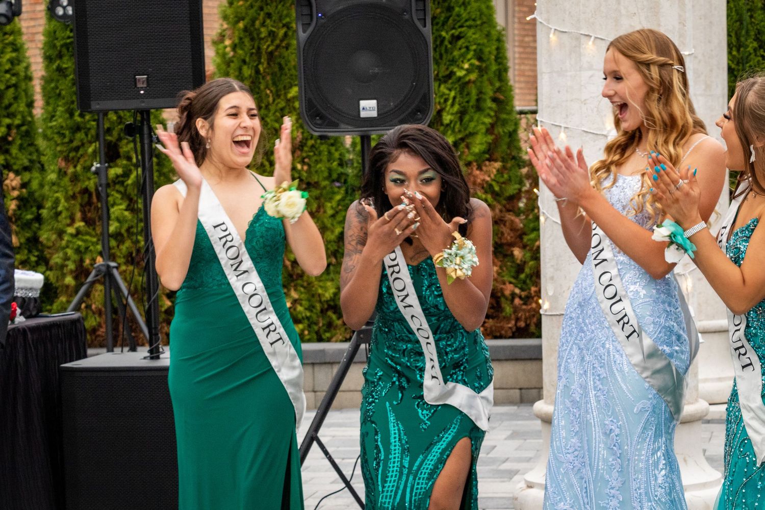 A group wearing formal dresses and sashes celebrate enthusiastically at an outdoor event.
