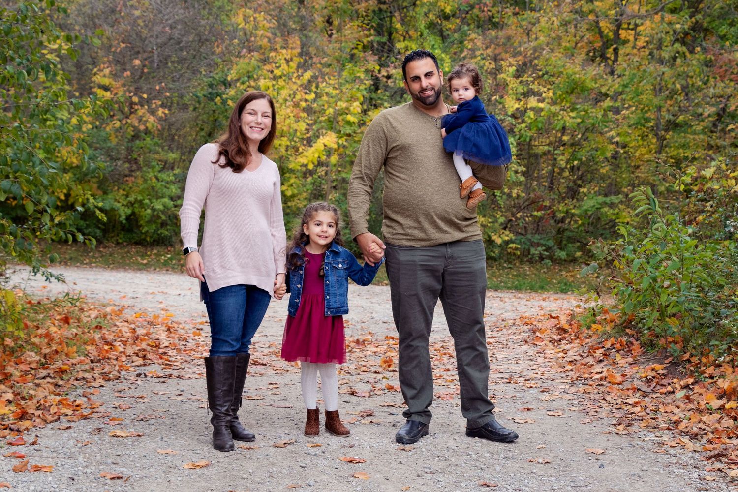 Family of four standing together on a leaf-covered autumn trail surrounded by colorful fall foliage.