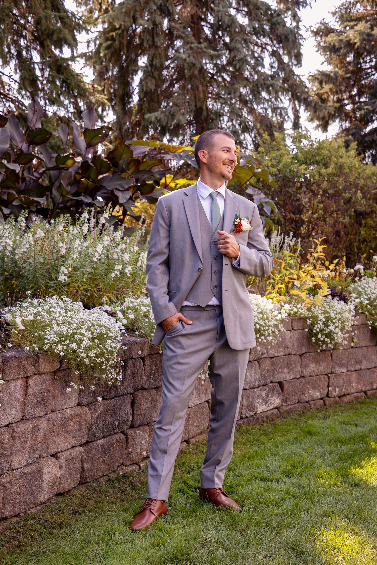 Groom in gray suit poses casually against rustic stone wall in garden setting.