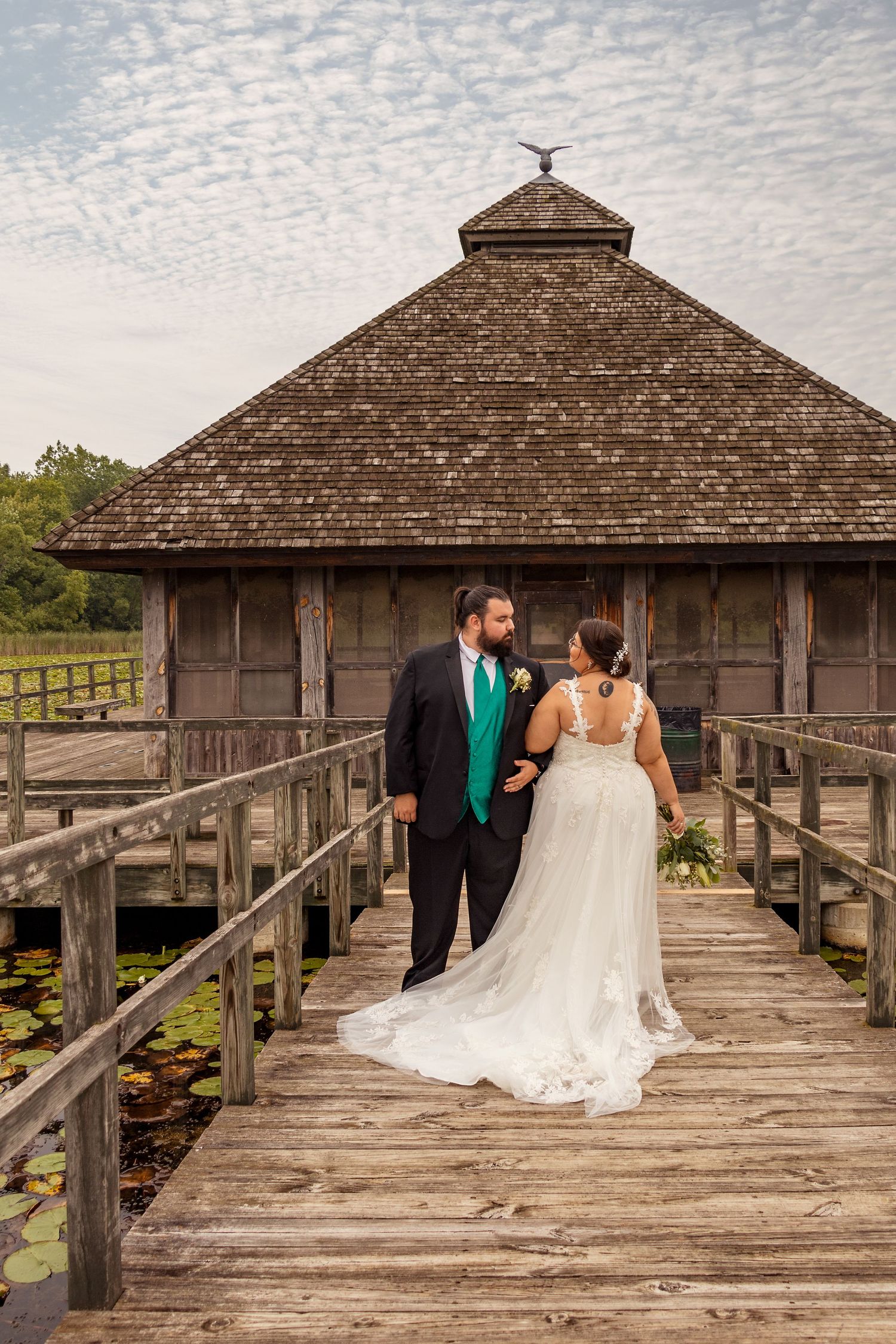A wedding couple poses on a wooden boardwalk leading to a rustic pavilion.