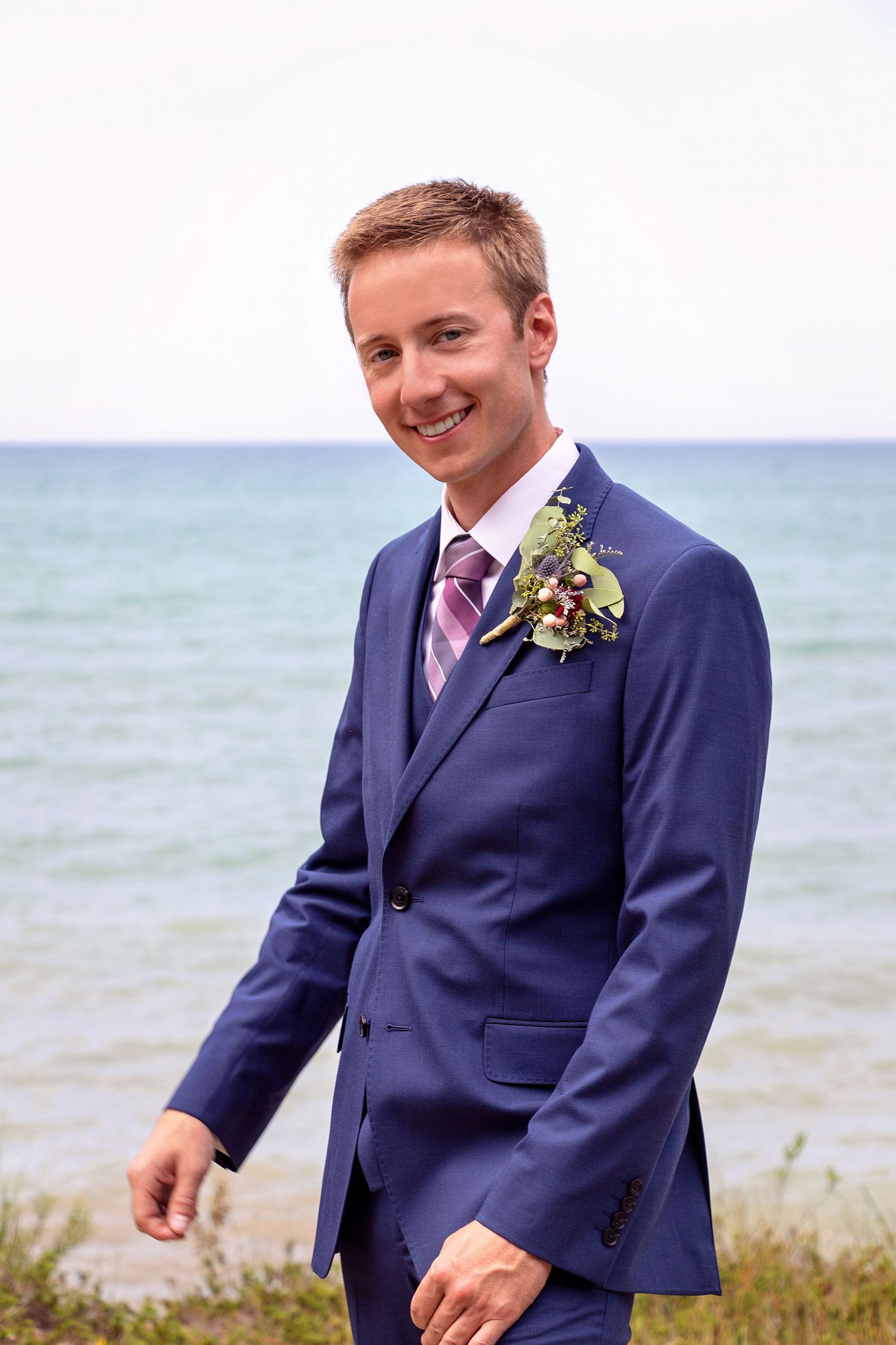 A groom in a navy suit with a pink tie and boutonniere smiles while standing near the ocean.