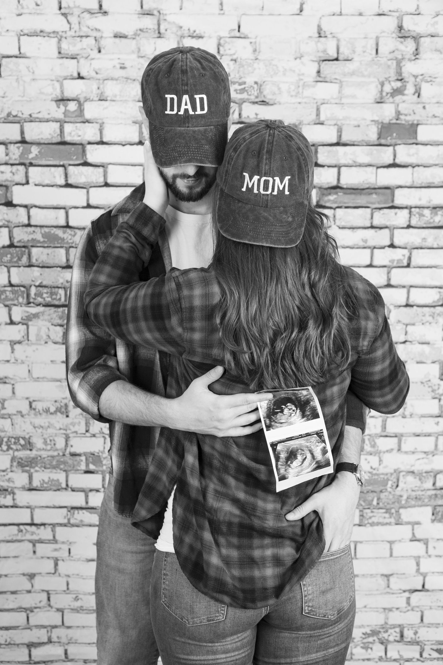 Black and white photos of a couple wearing Dad and Mom hats posing against a brick wall.