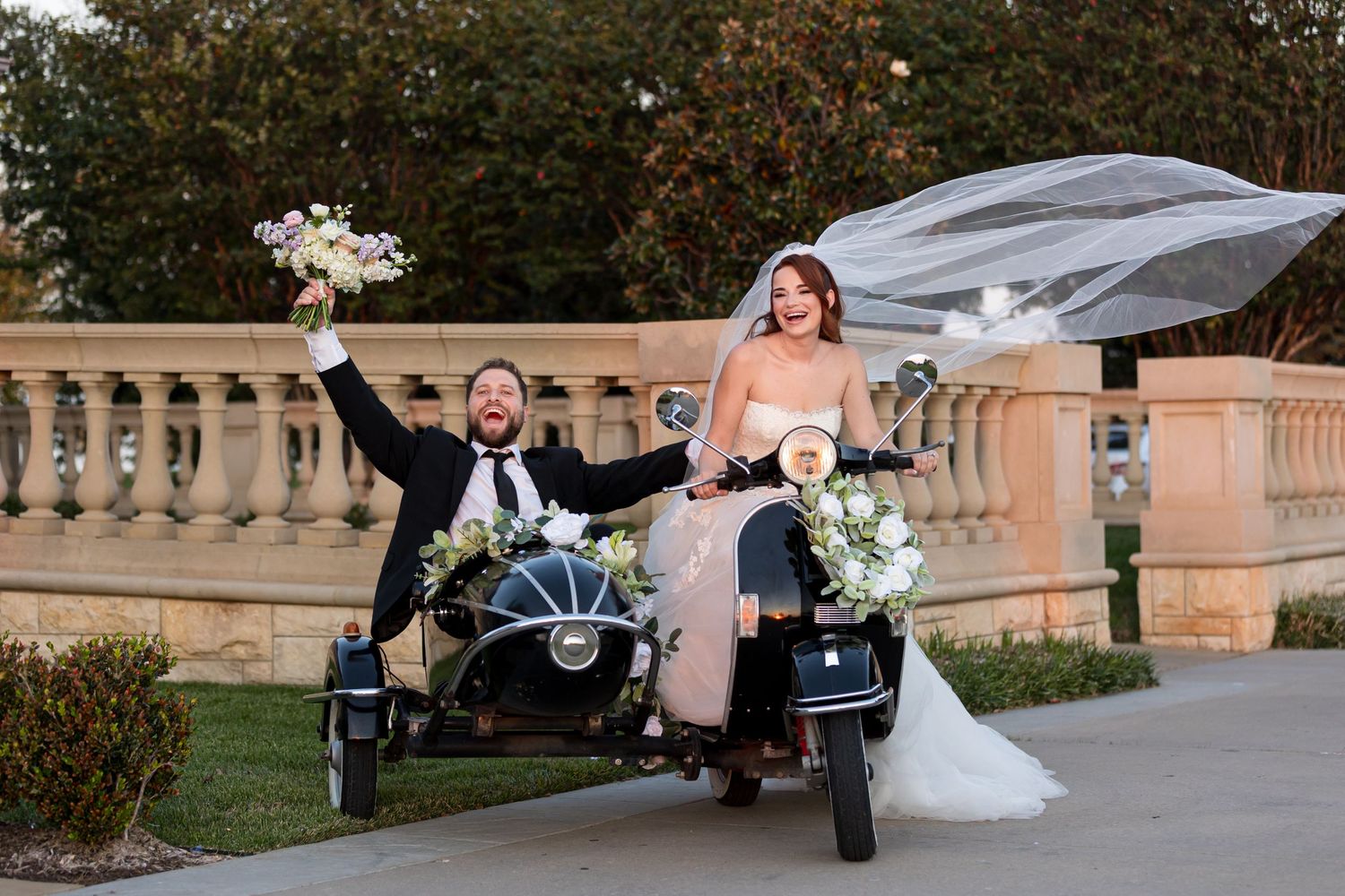 bride and groom on black vintage vespa with side car, veil blowing, groom raising flowers, lot of fun, off we go from the Olana