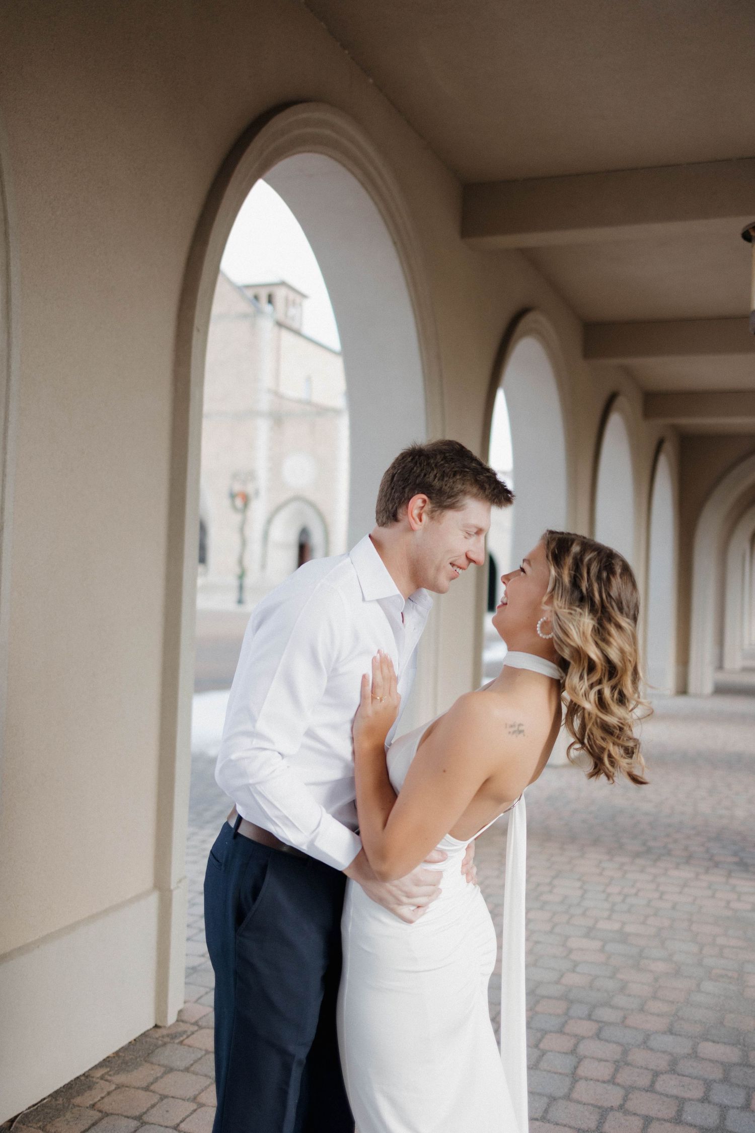 Wedding couple embraces intimately under a covered walkway with arched columns.