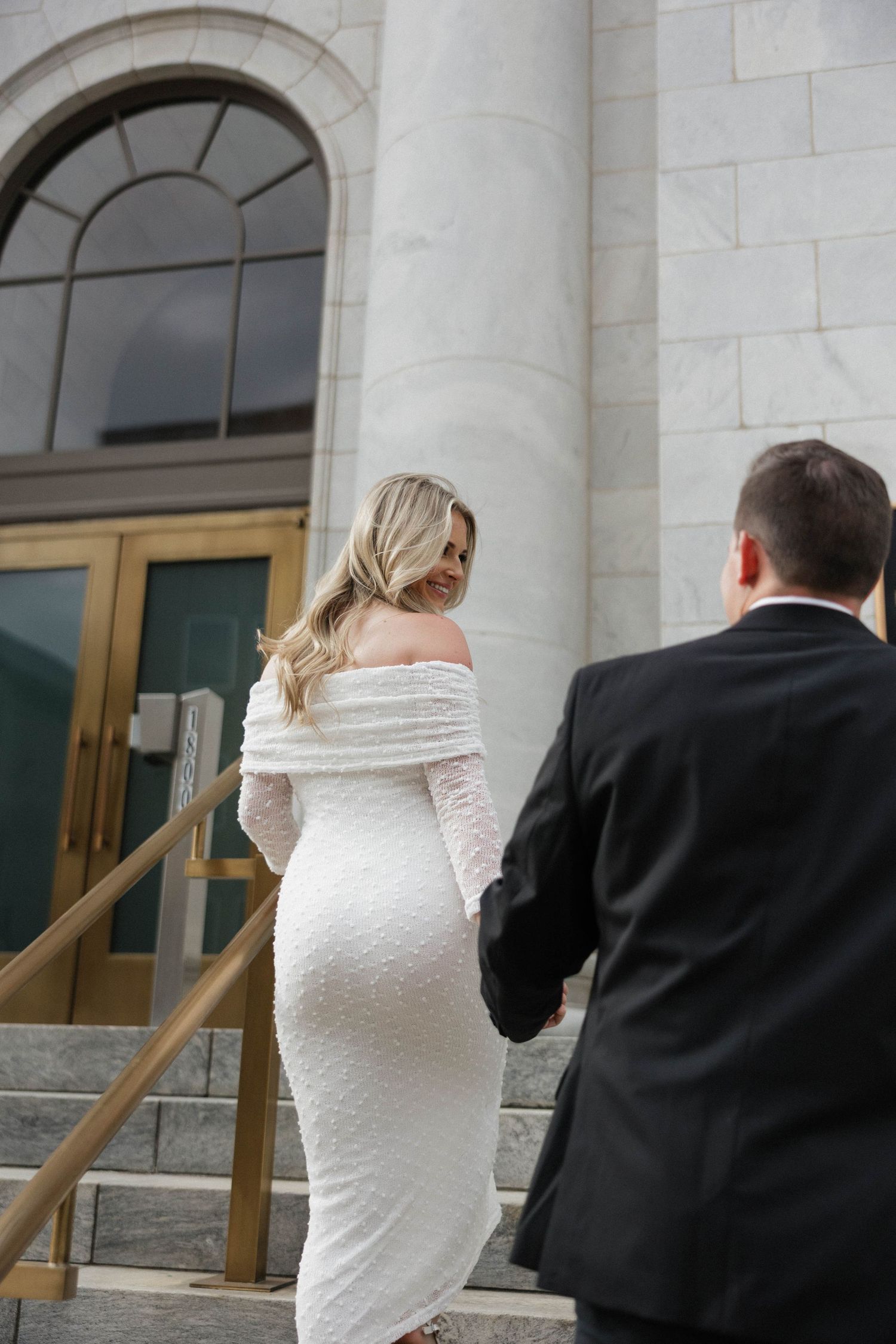 Wedding couple walks hand in hand along columned building with brass handrails.