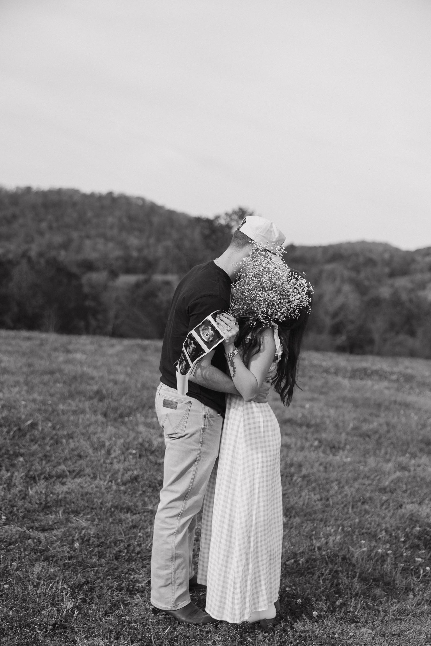 Black and white photo of a couple kissing, holding ultrasound photos and a bouquet, announcing pregnancy outdoors.