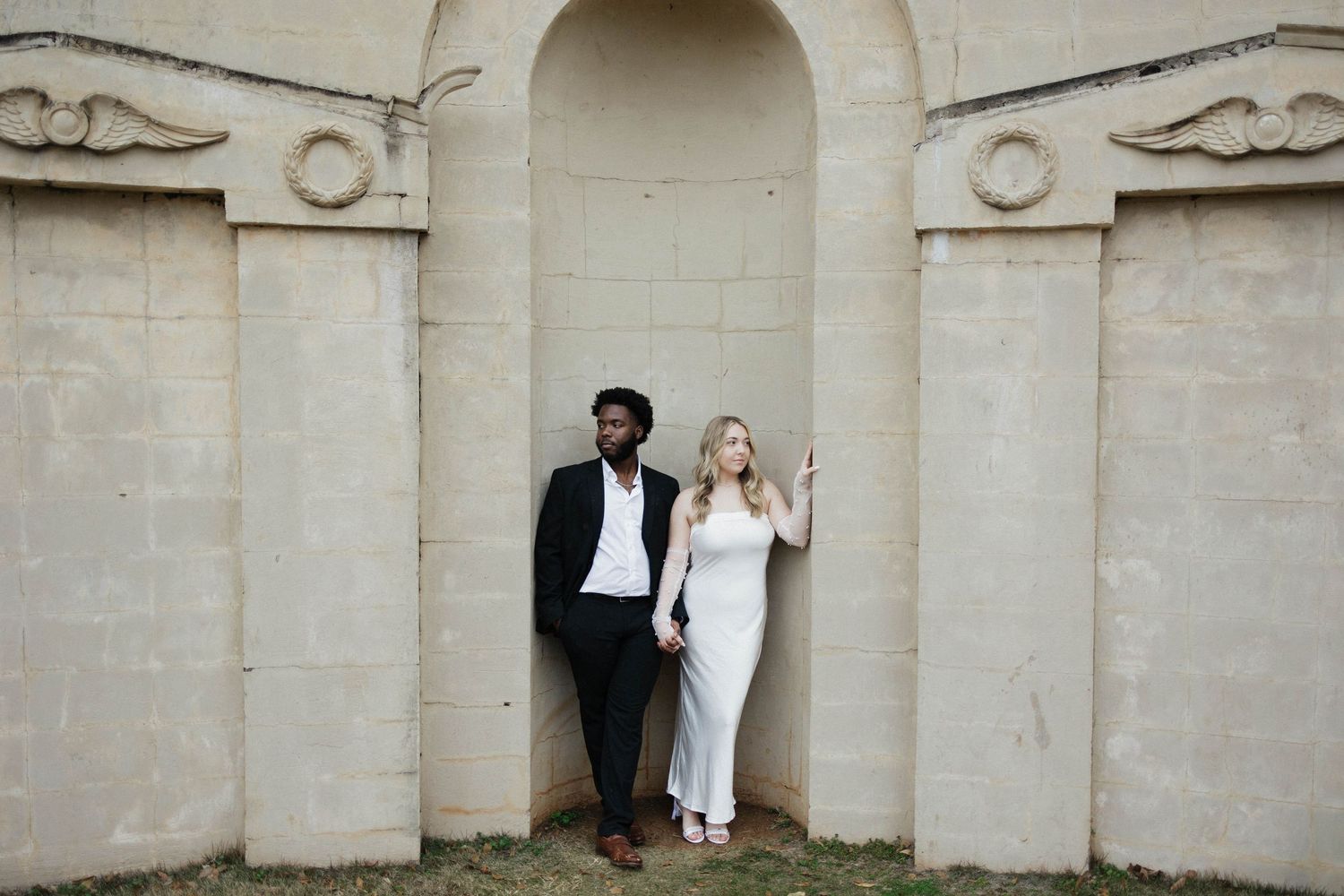 Wedding couple poses against stone wall with architectural details.