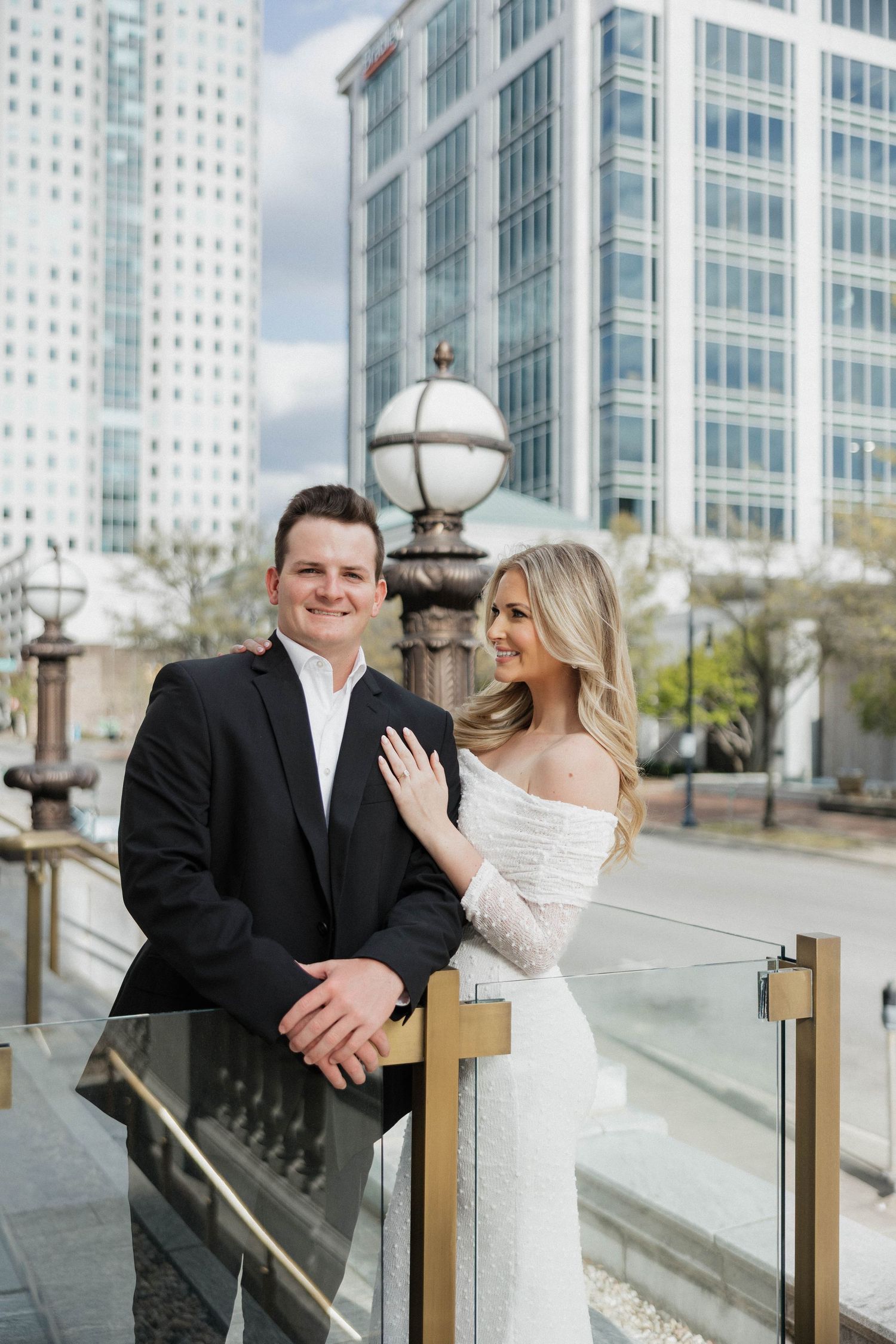 A couple in wedding attire pose together against a modern city backdrop with glass buildings.
