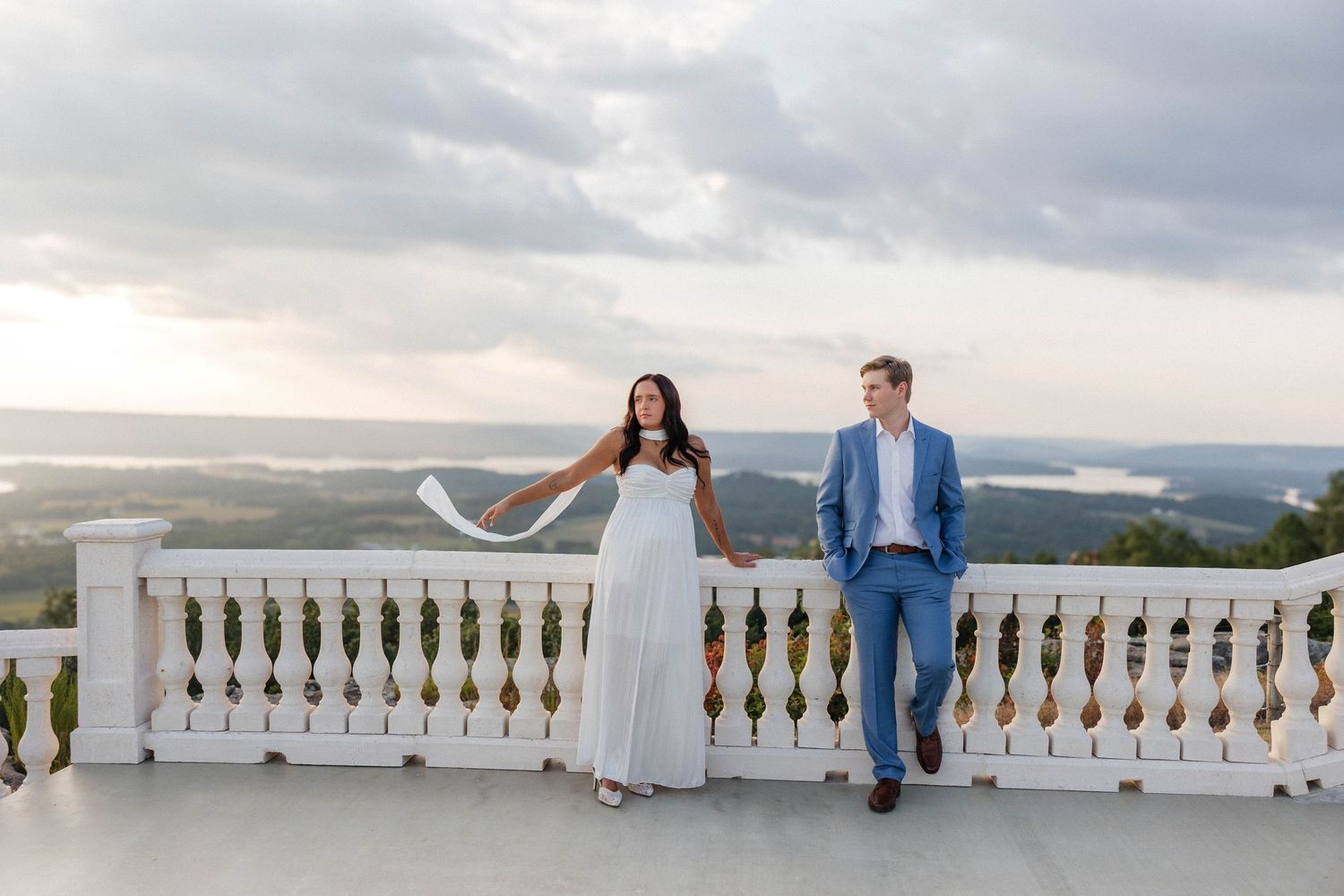 Romantic couple sitting on white stone balustrade with scenic mountain backdrop.