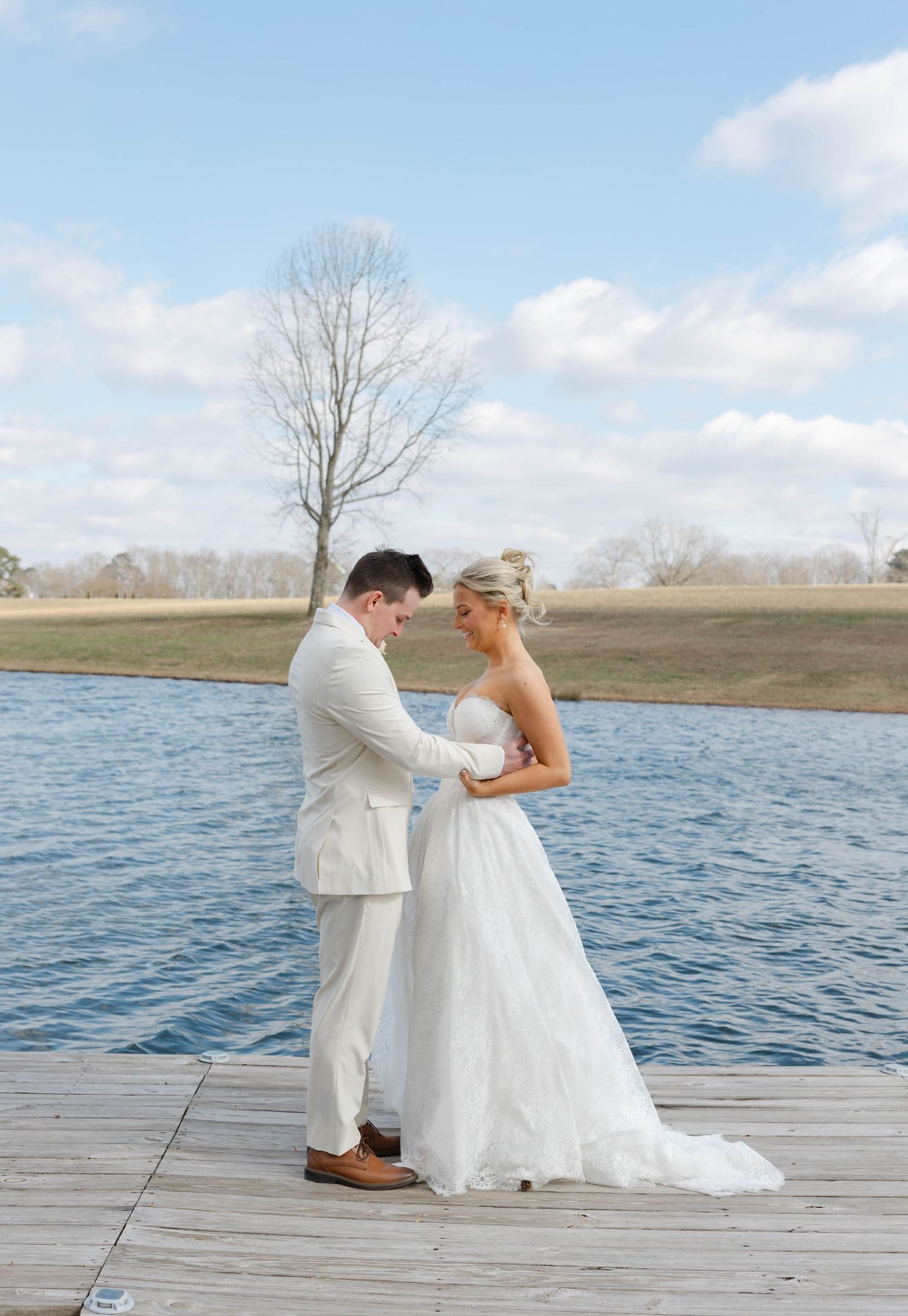 Bride and groom holding hands on a lakeside dock, wearing white wedding attire, with a serene blue sky backdrop.