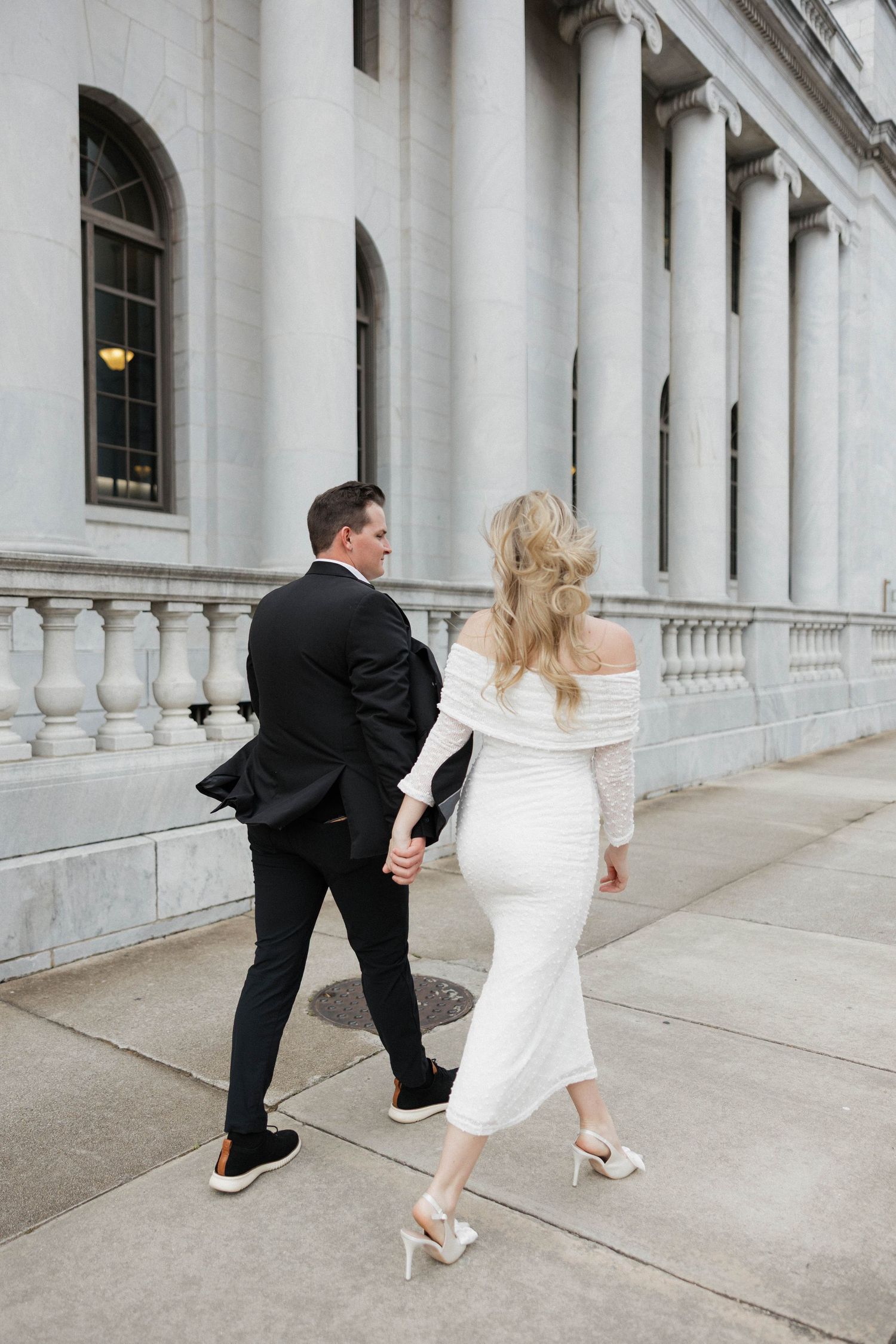 Playful couple in wedding attire dance and twirl on a city sidewalk.