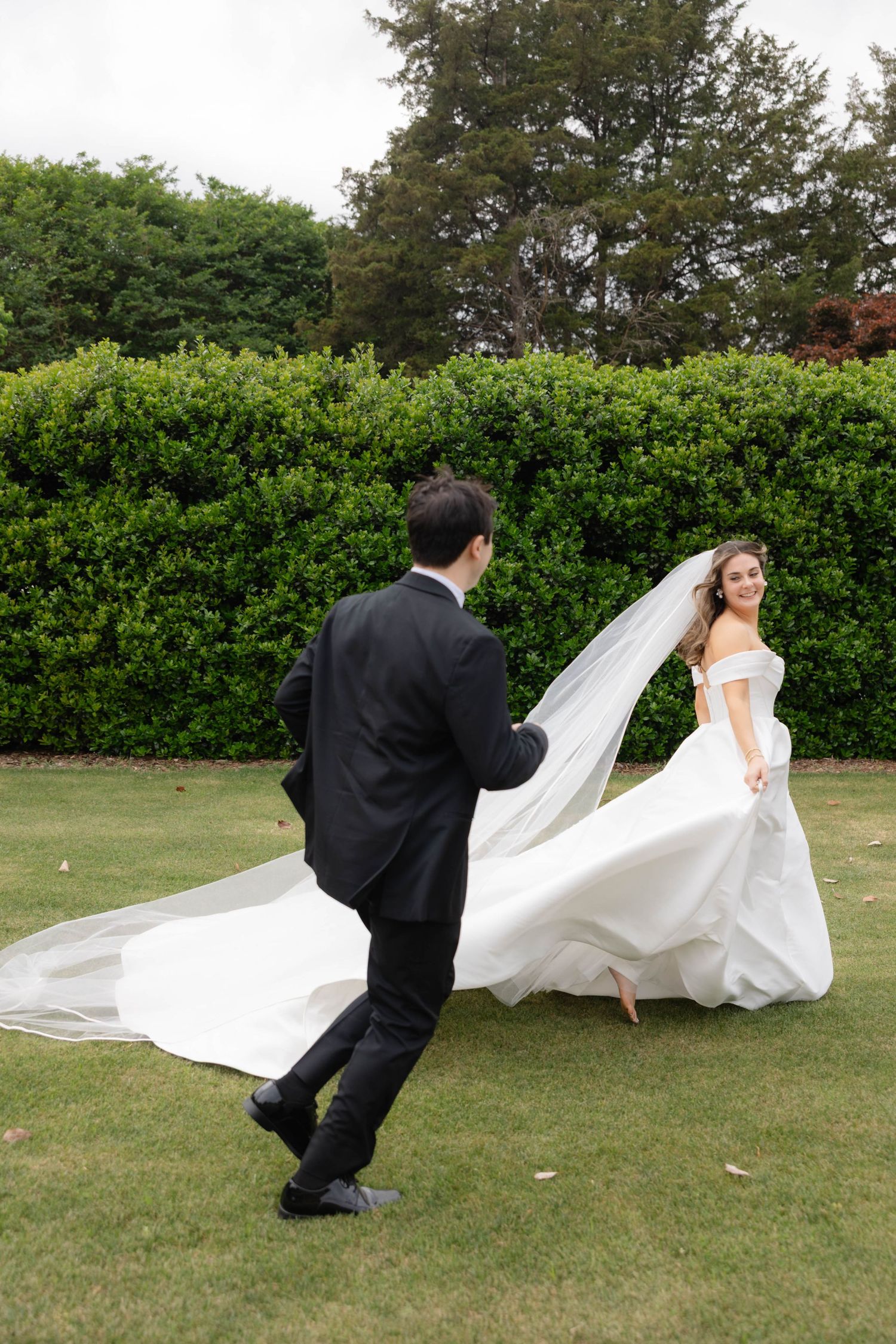 Bride in flowing white gown and veil twirls joyfully as groom in black tuxedo chases her across lush garden lawn.