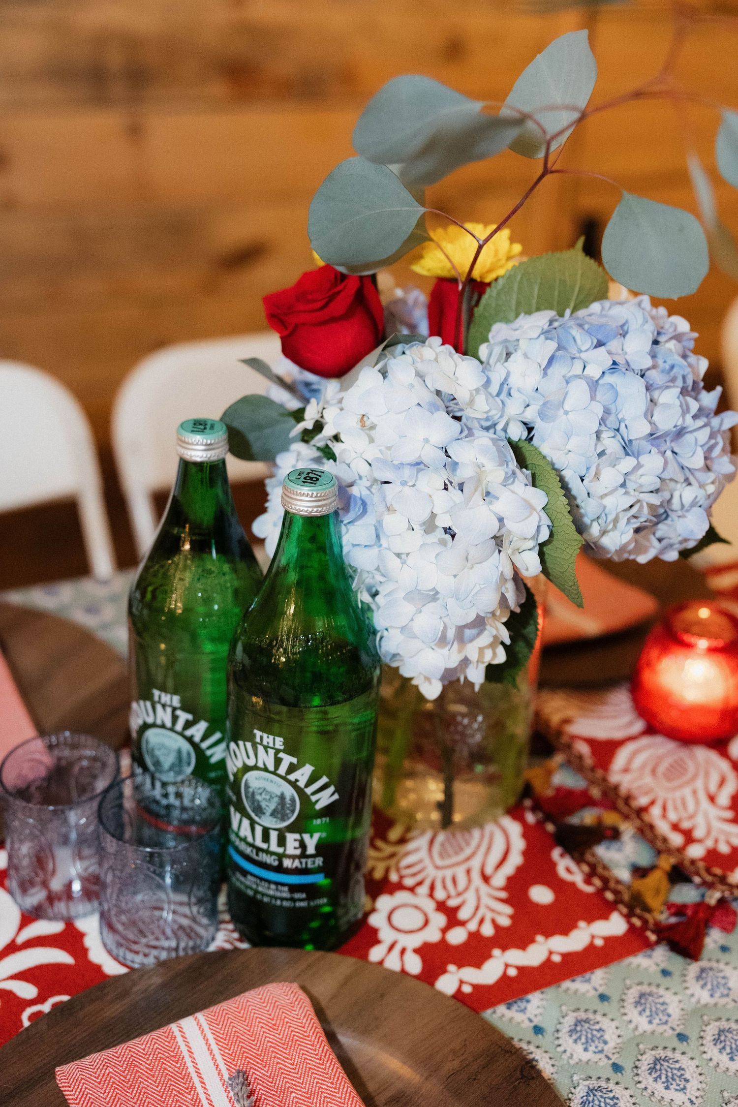 Wedding reception table decor with green glass bottles, white hydrangeas, and red accents on a rustic wooden surface.