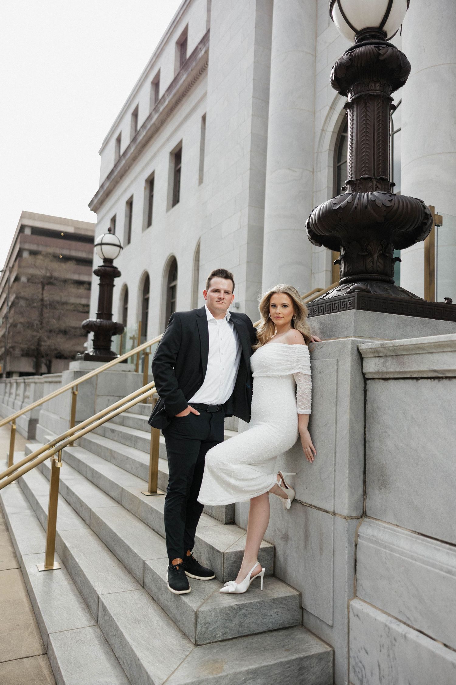 Couple in wedding attire poses playfully on grand stone steps with vintage lamp posts.