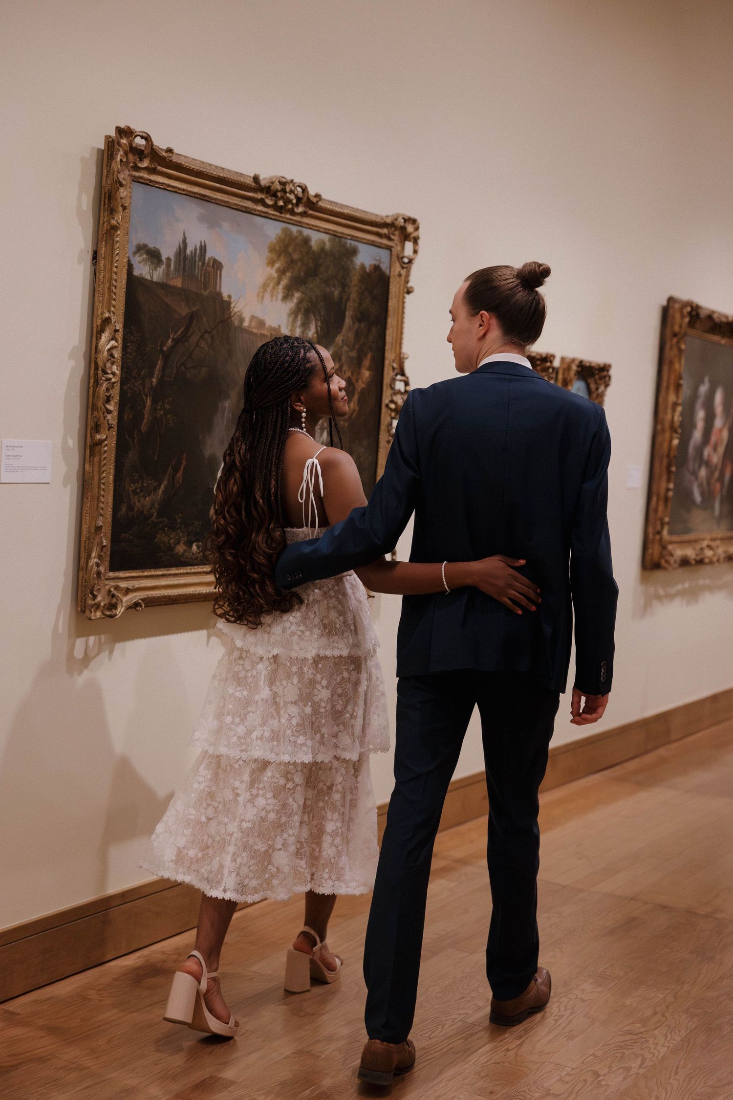 A couple views artwork in an ornate gallery while dressed in formal attire.
