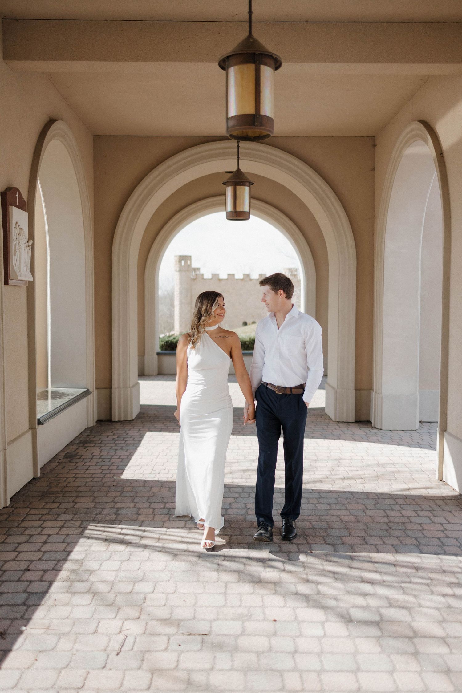 Couple walks together through architectural colonnade with arched windows.