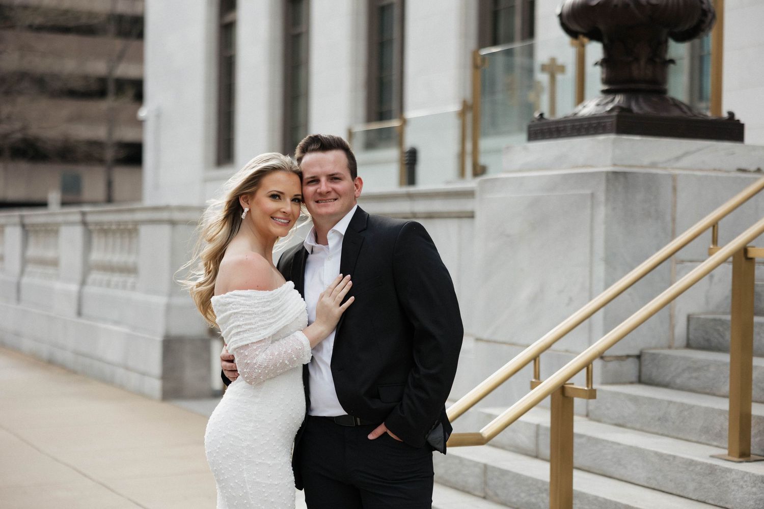 Romantic couple in wedding attire poses on stone steps of a grand building.