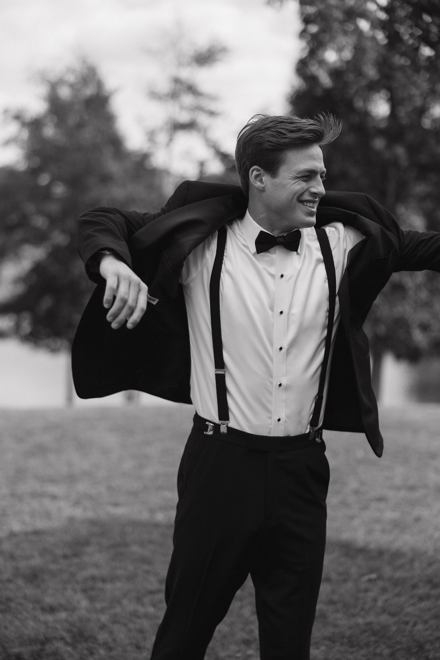 Groom in suspenders and formal wear poses playfully outdoors.