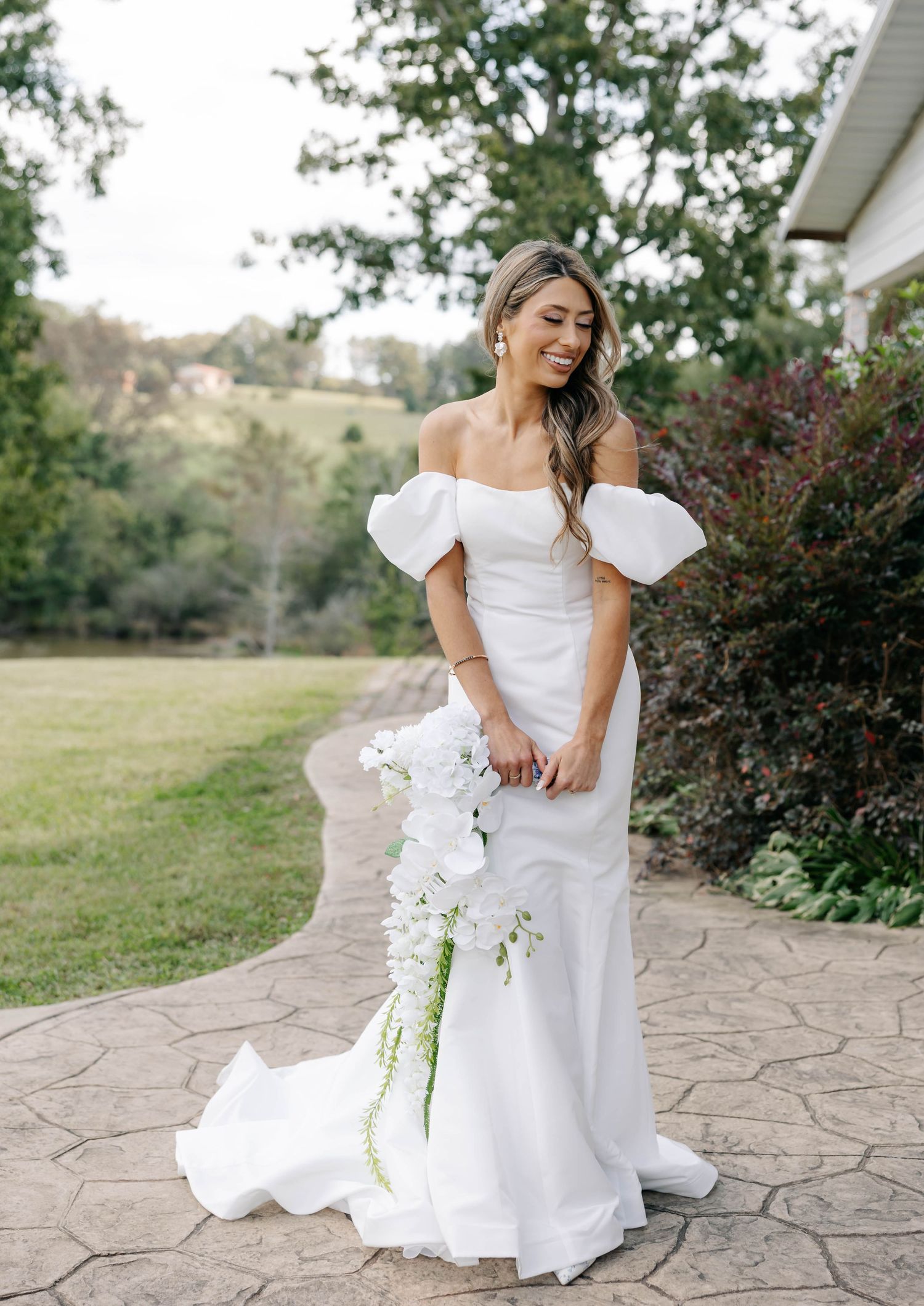 Bride in elegant white dress poses on stone patio with garden backdrop.