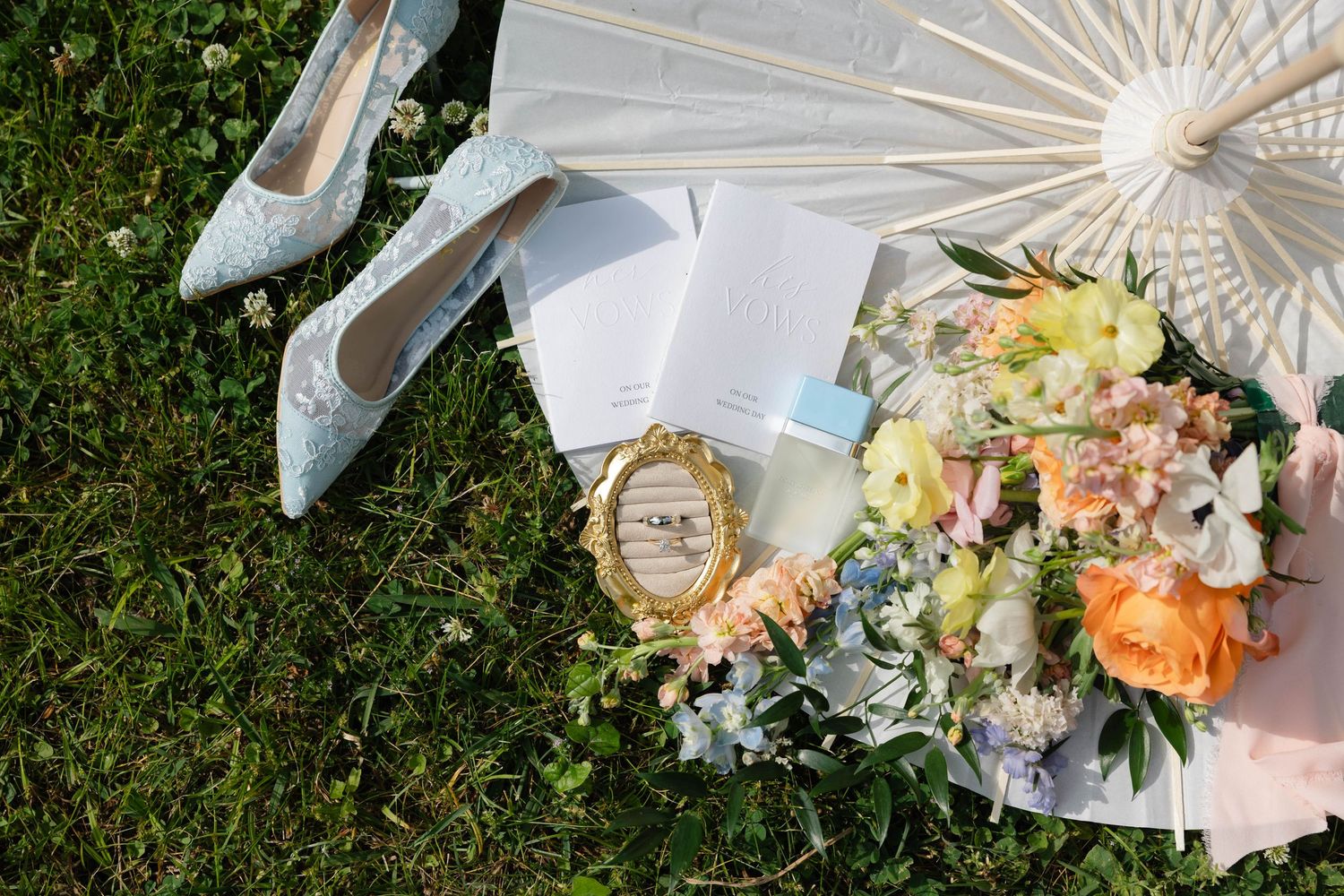 Wedding flat lay with lace heels, floral bouquet, parasol, invitations, and gold ring box on green grass.