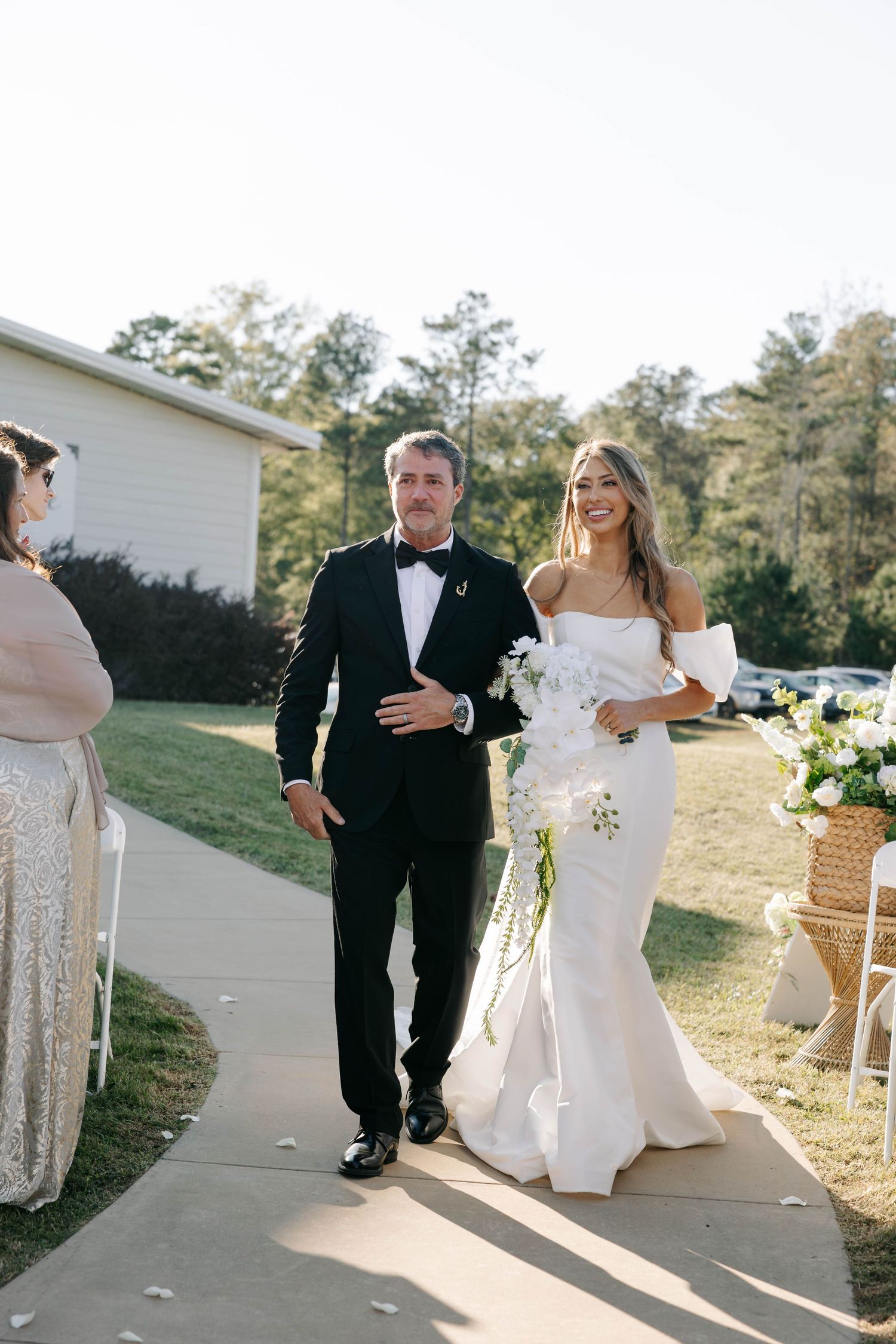 Wedding couple walks along a sunlit path outside a white venue building.
