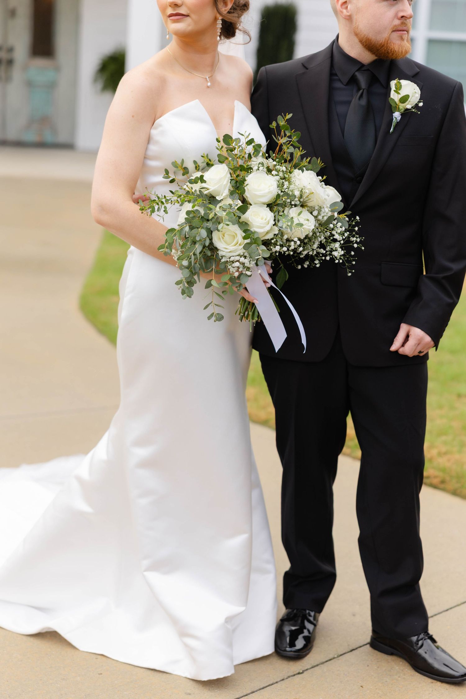 Bride in elegant white gown standing with groom in black suit holding white bouquet.