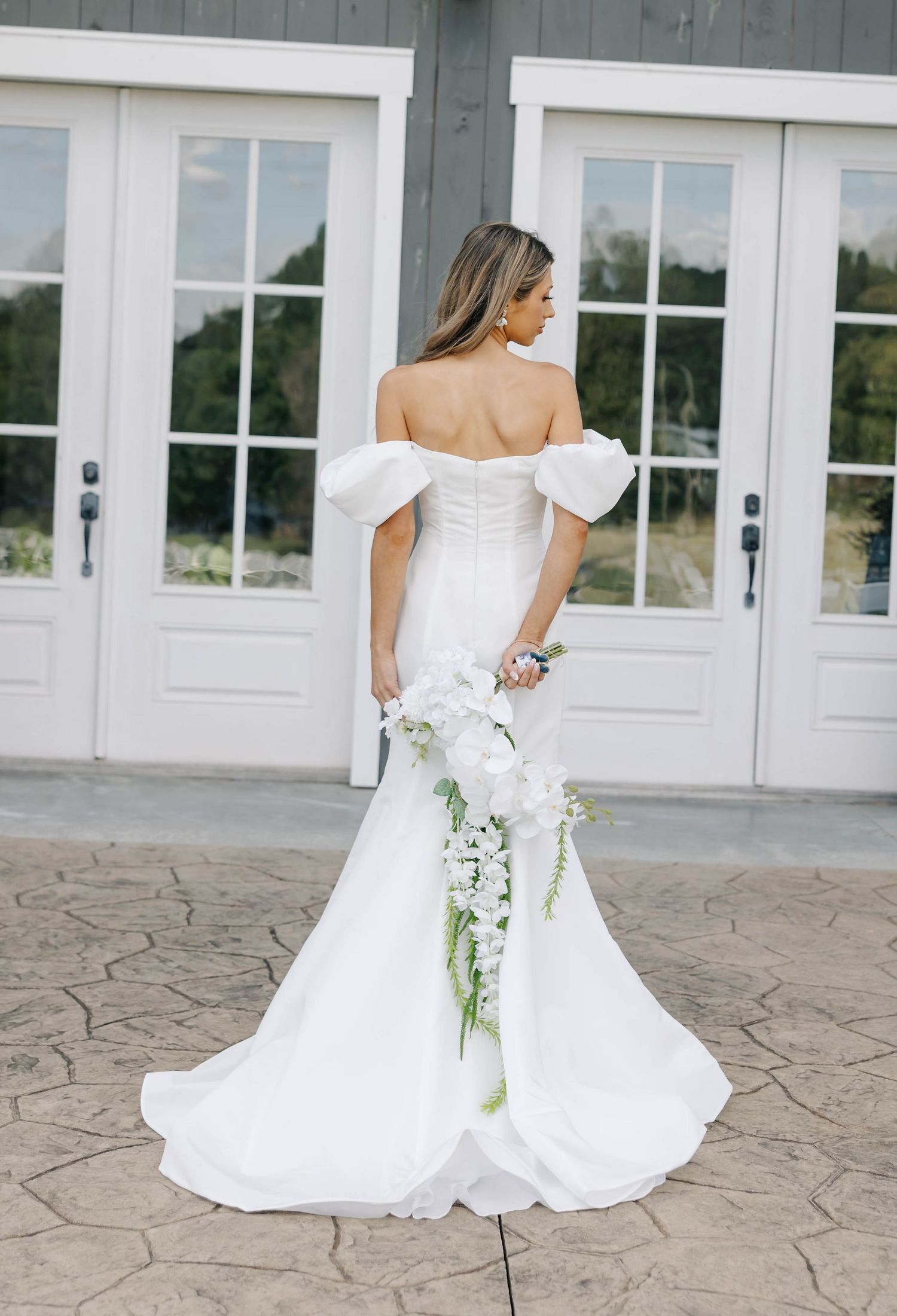 Elegant white wedding dress flows across stone patio in front of glass doors.