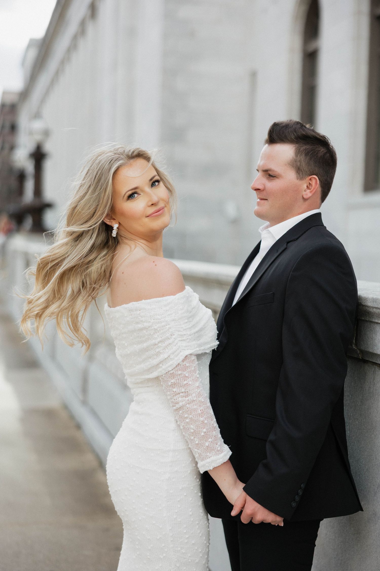 A bride in white lace and groom in black suit pose against a classical stone building.