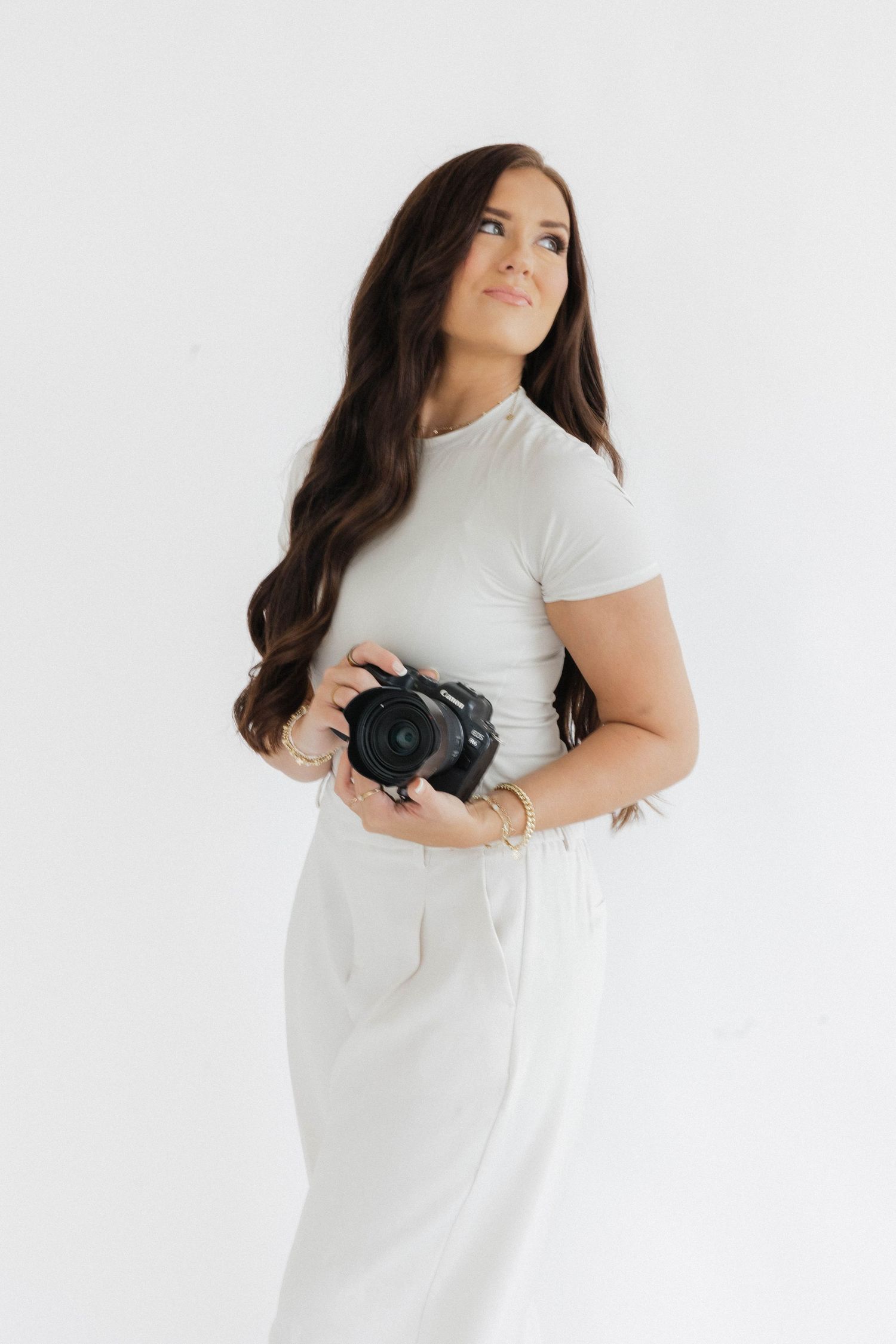 Professional photographer in white dress poses with camera against minimal background.