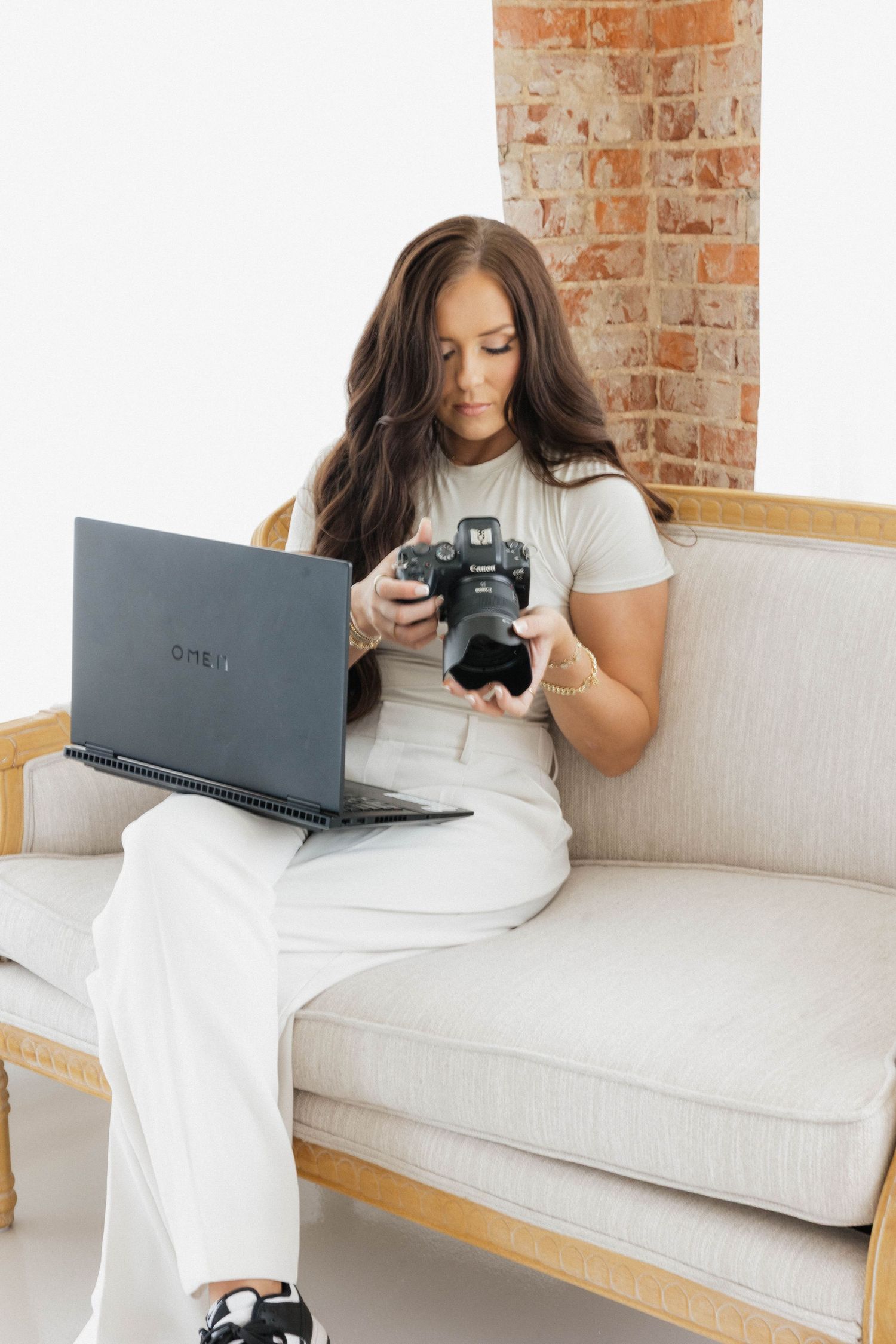 Person in white outfit reviewing photos on camera and laptop while sitting on beige couch.