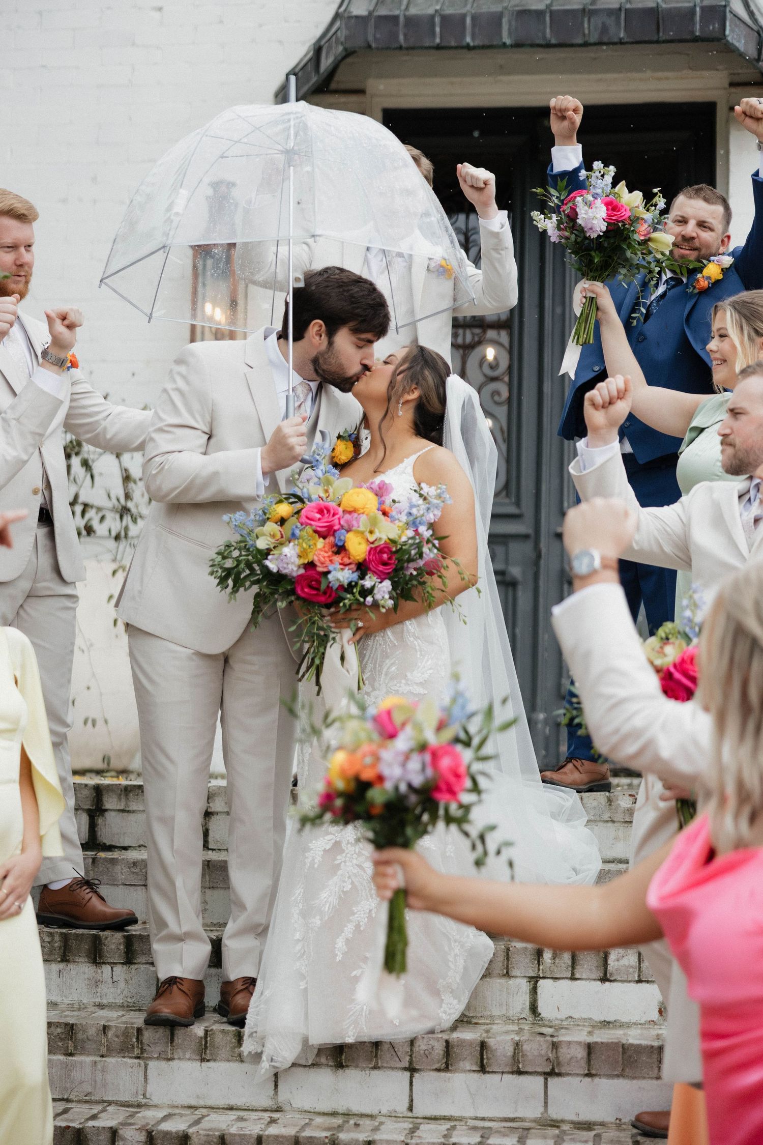 Wedding party celebrates with raised arms and umbrellas in front of a stone wall.