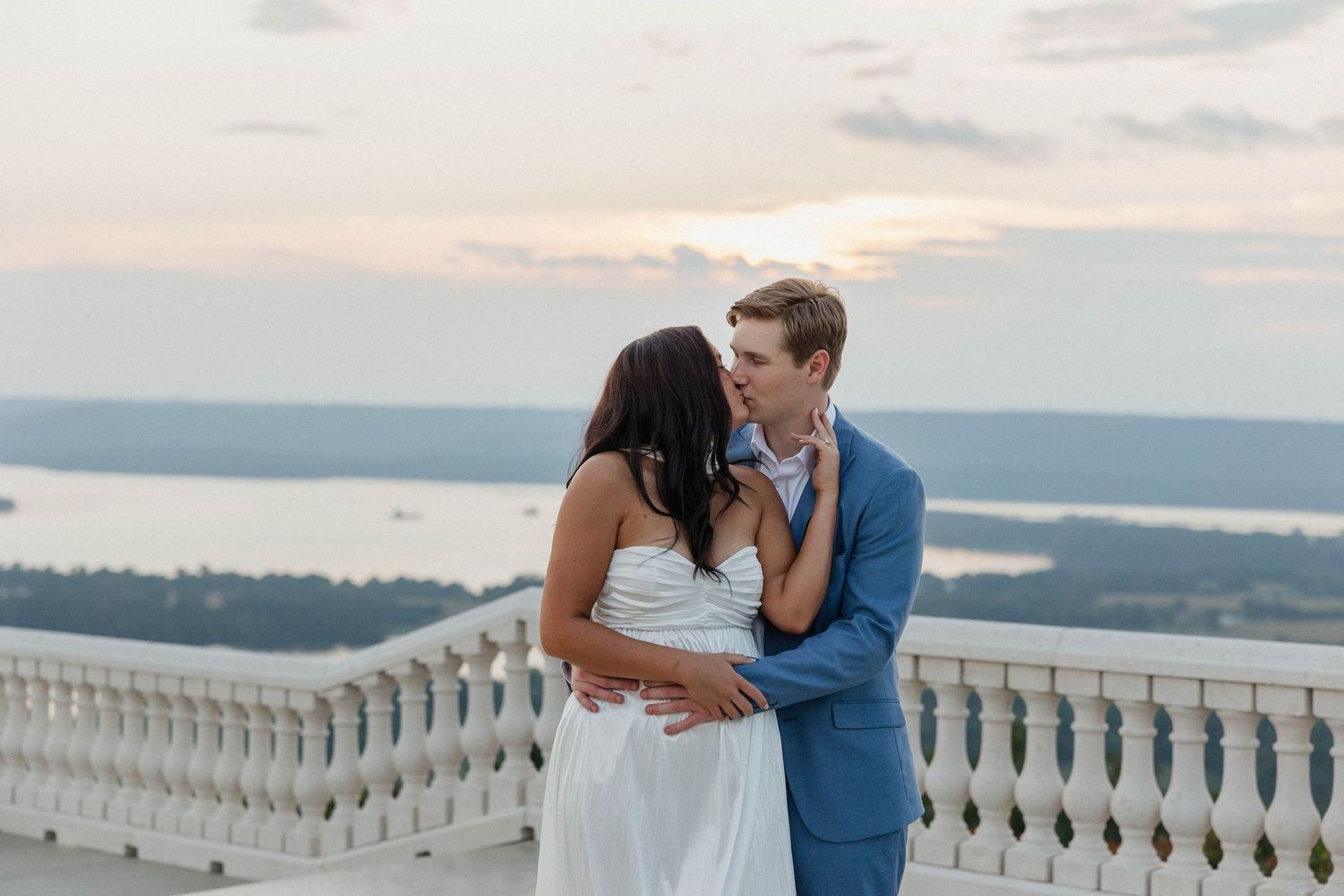 Elegant couple in formal attire sharing an intimate moment on a balcony with lake views.