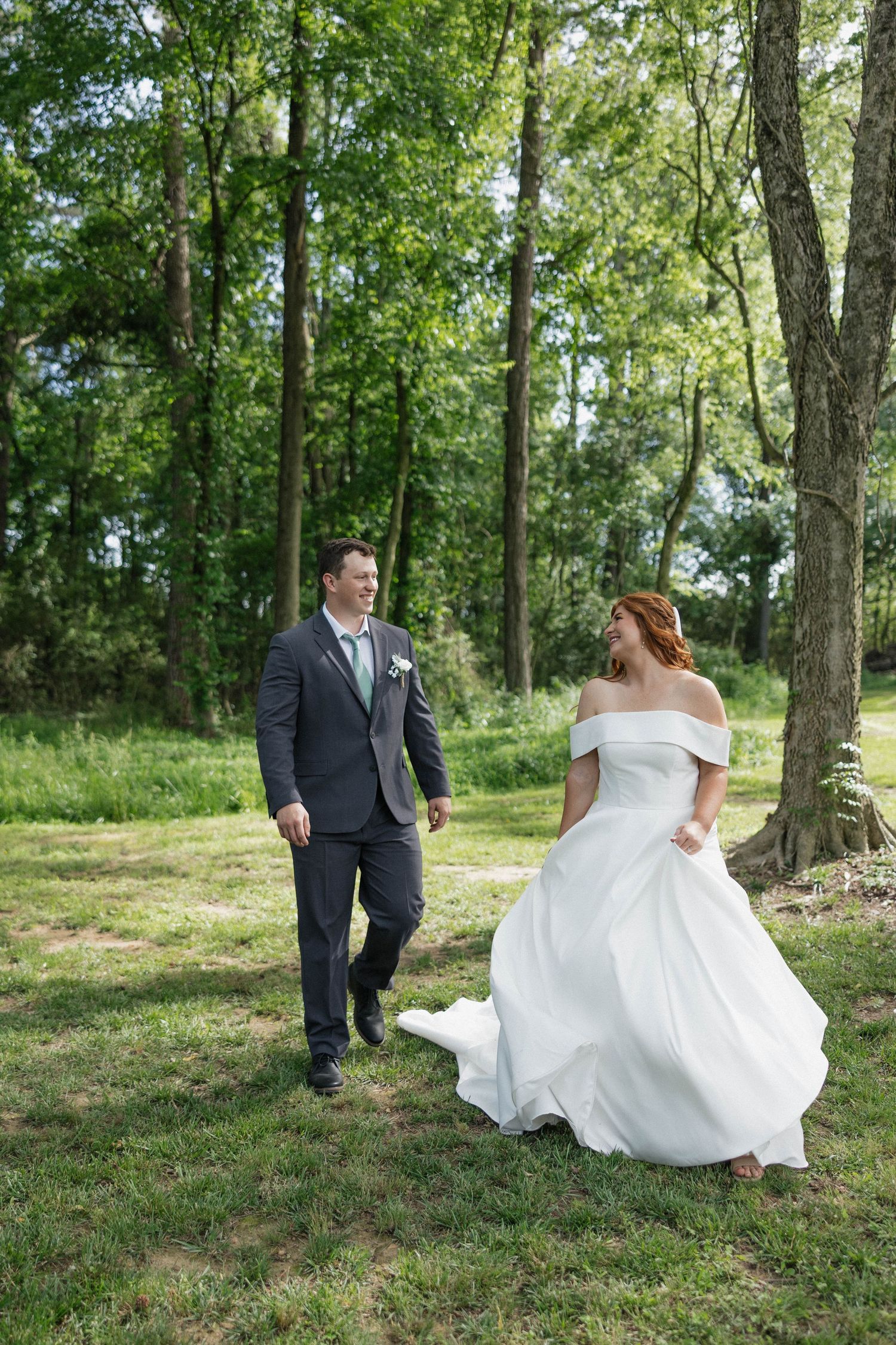 A couple in wedding attire playfully twirls in a sunlit wooded area during their photo session.