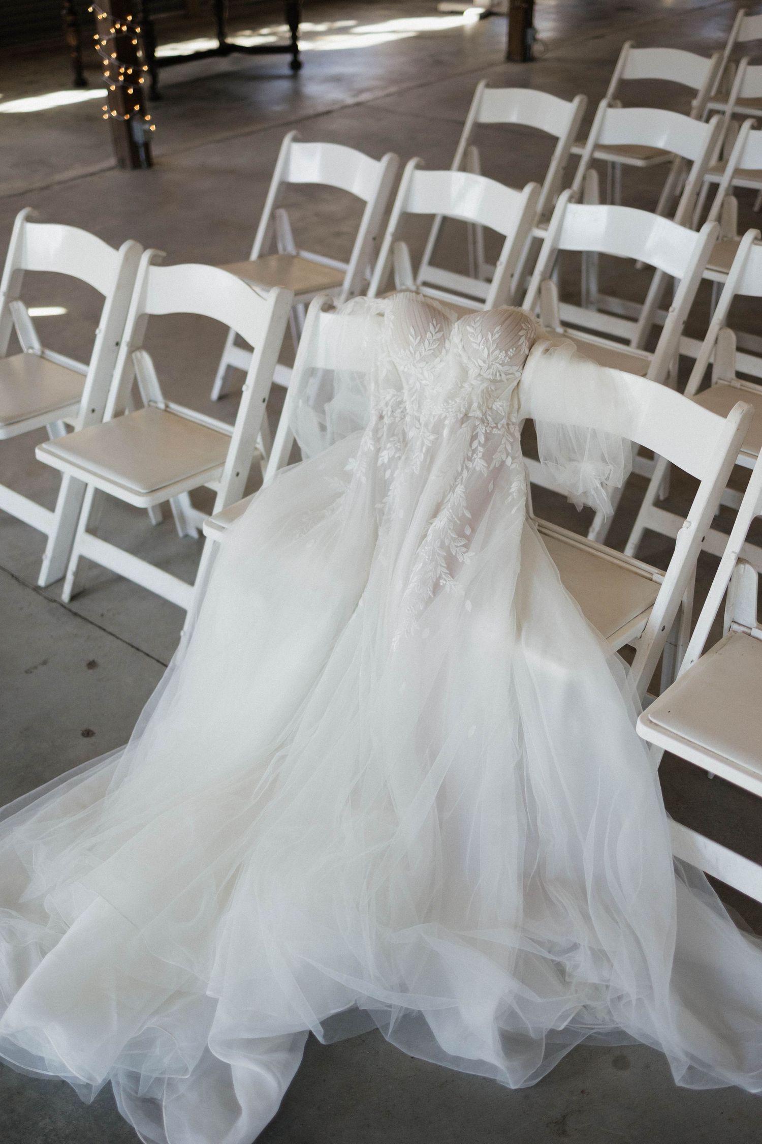 Elegant white wedding dress draped over folding chairs in ceremony venue.