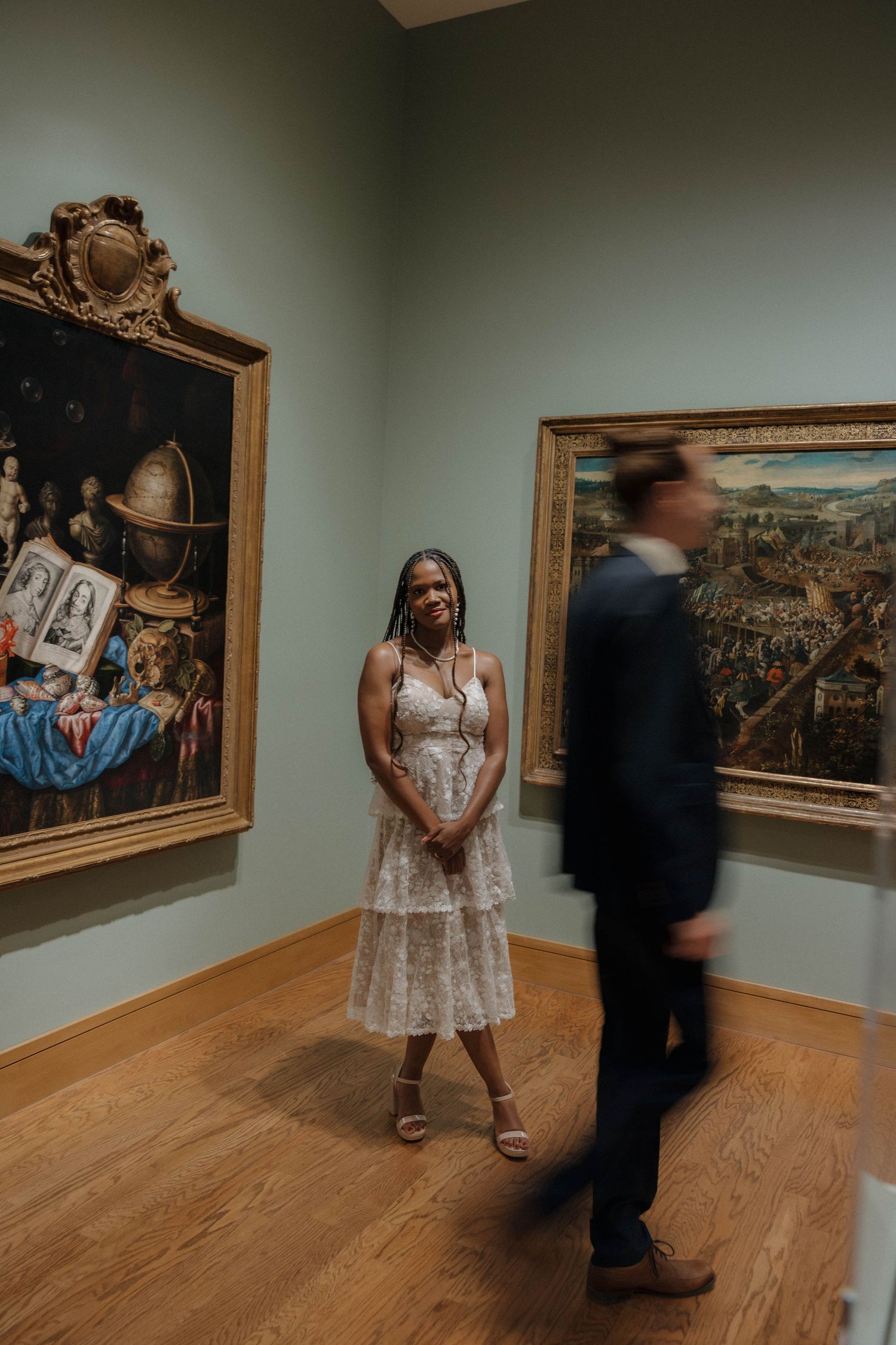 A couple in wedding attire standing in an art museum gallery surrounded by classical paintings.