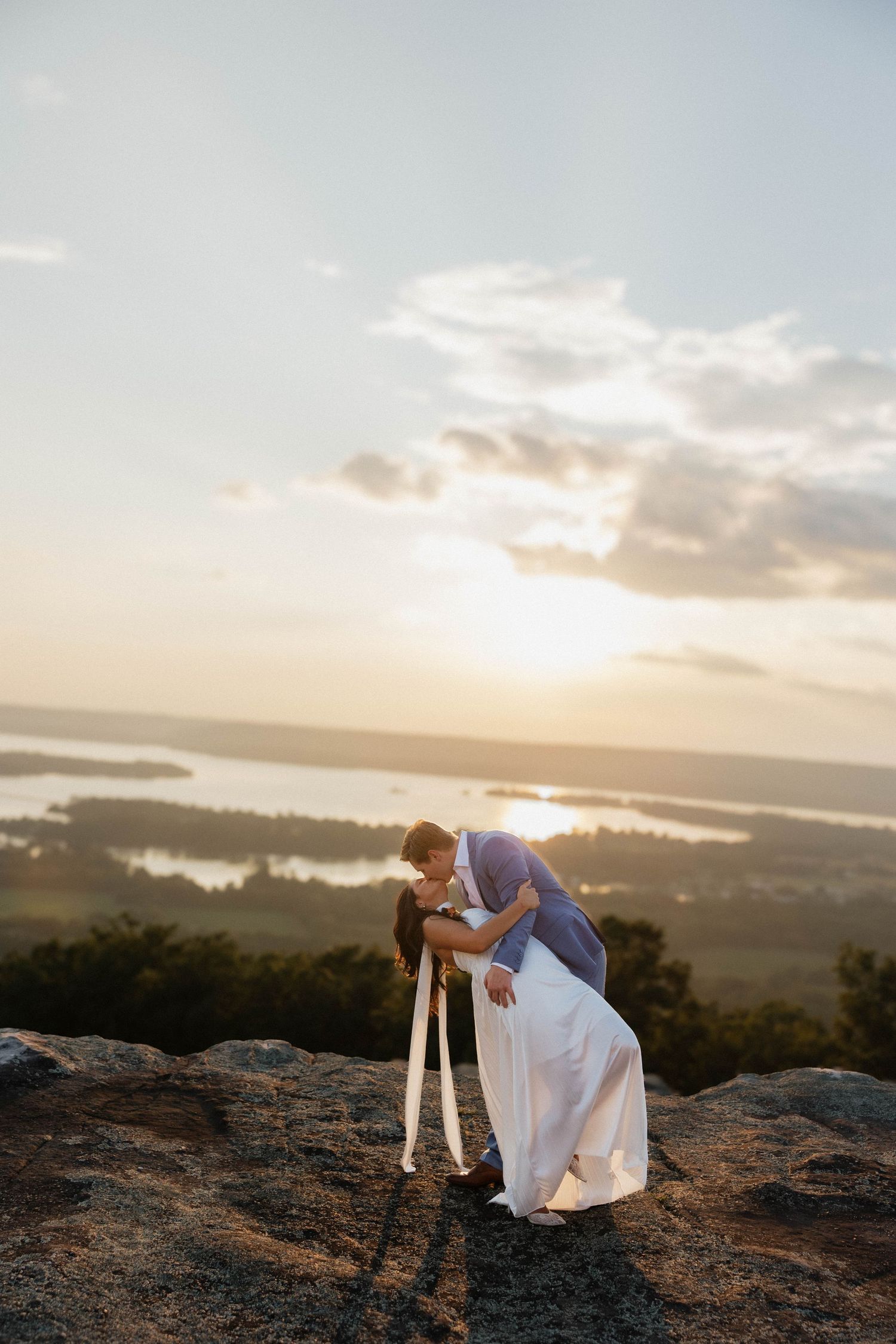 Couple in wedding attire on rocky overlook during golden hour.