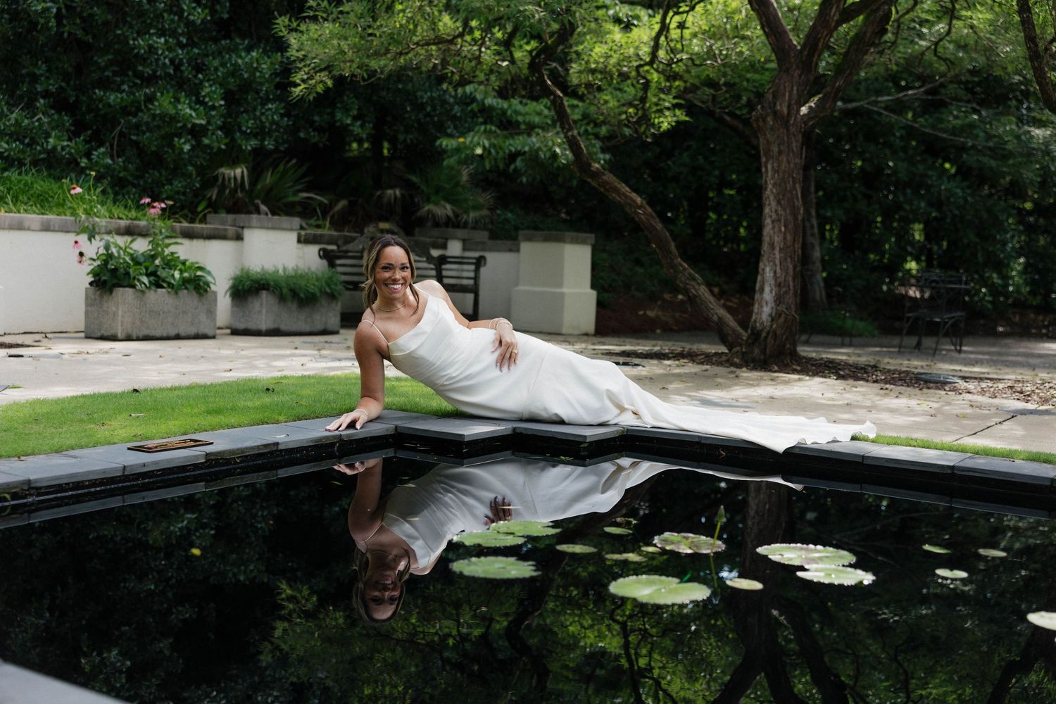 Person in white dress leaning over dark pond with lily pads in garden setting.