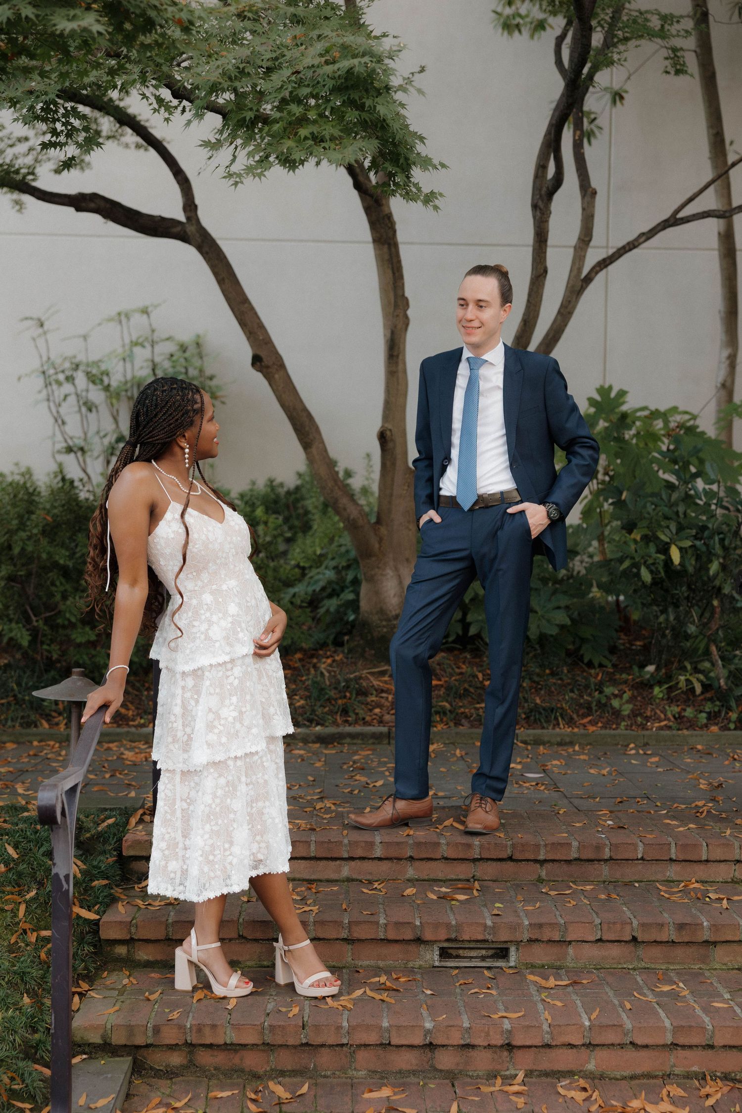 Stylish couple poses on brick steps under trees in formal attire.