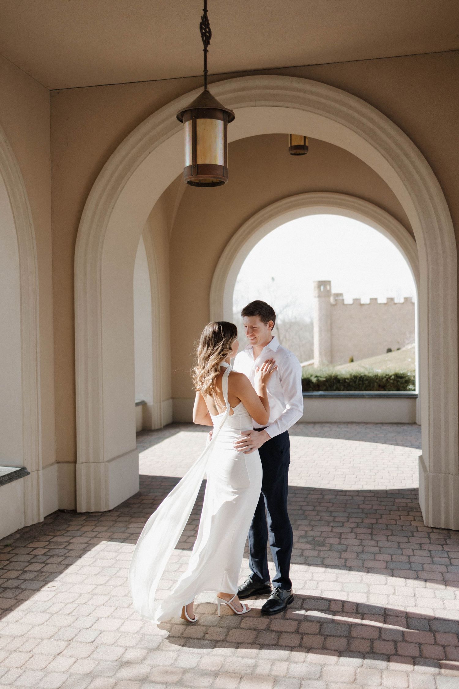 Romantic couple shares intimate moment under arched colonnade with castle view.