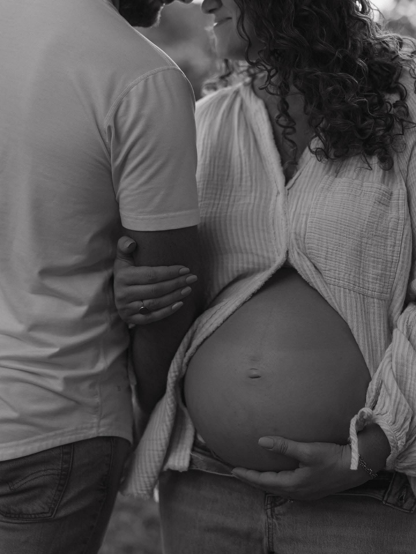 Black and white maternity photo of a couple embracing, showcasing a pregnant woman's bare baby bump outdoors.