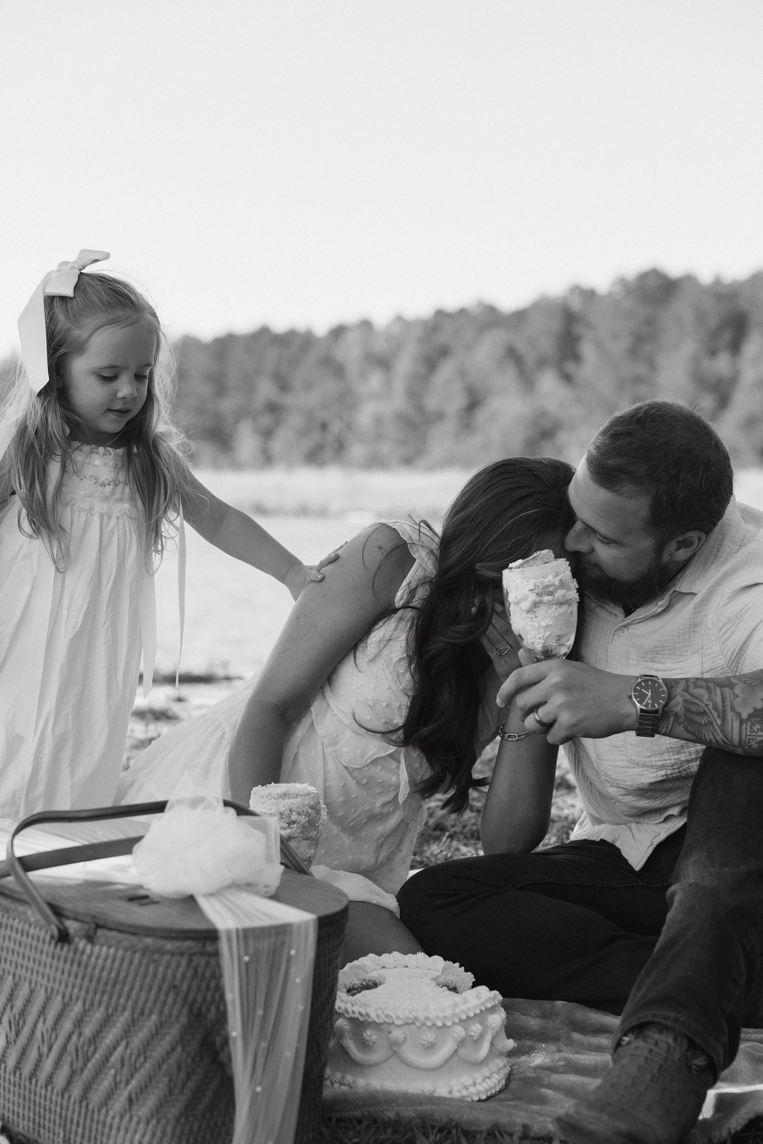 Black and white photo of a family enjoying a picnic by a lake, with a little girl in a bow watching her parents share a treat.