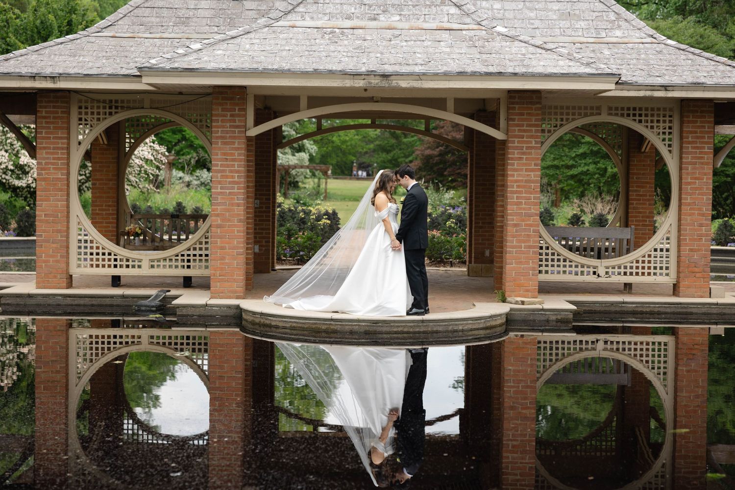 Bride and groom share a romantic moment in an elegant brick gazebo reflected in still water at a lush garden wedding.