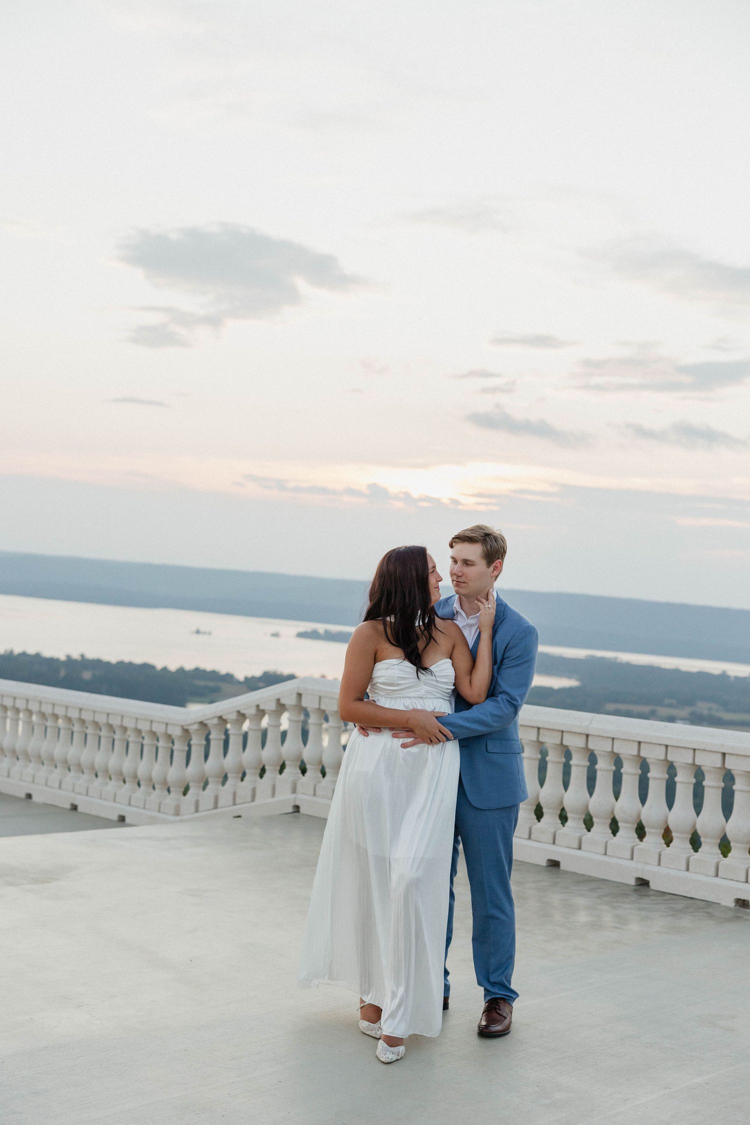 A romantic couple embraces on a balcony overlooking a scenic lake view at sunset.