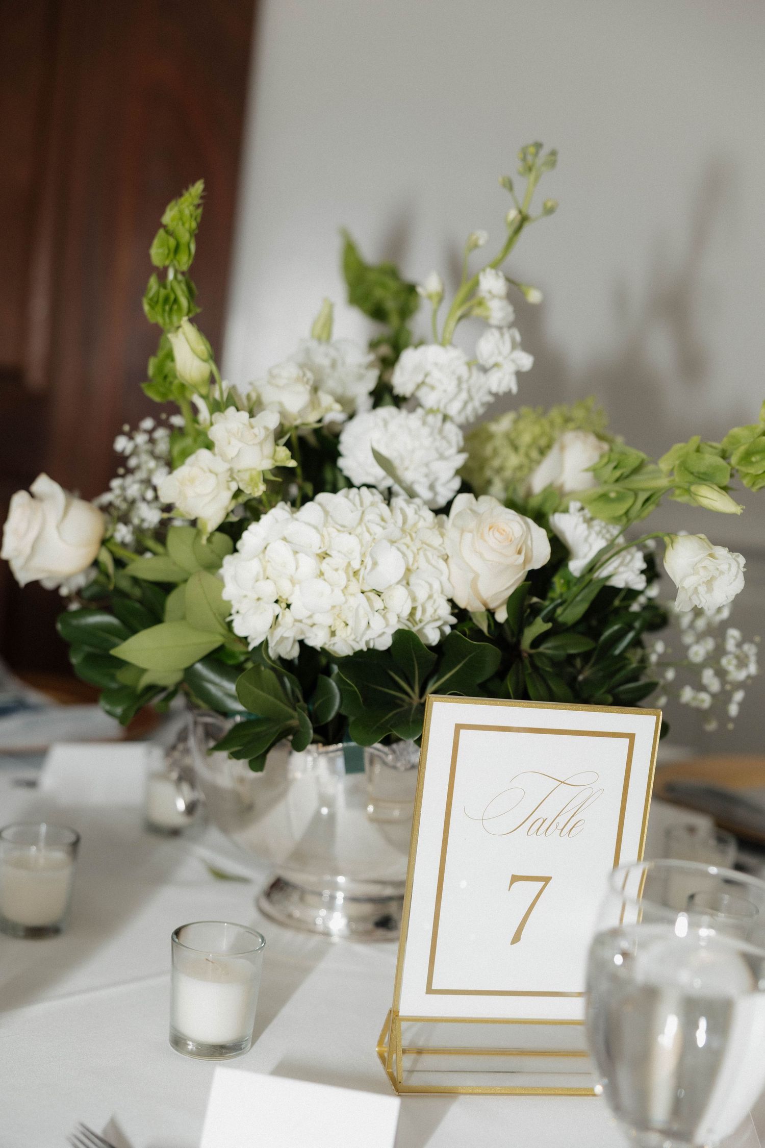 Elegant white floral centerpieces and table numbers in gold frames adorn wedding reception tables.