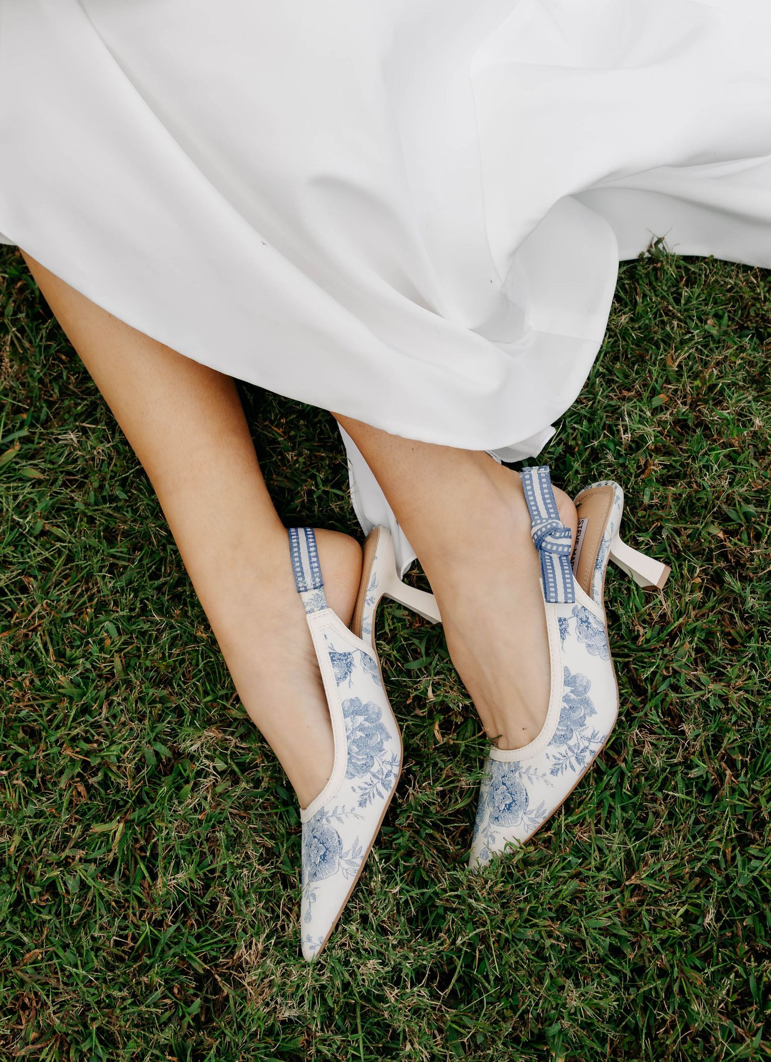 Delicate blue and white floral heels peek out from beneath a flowing white wedding dress on grass.