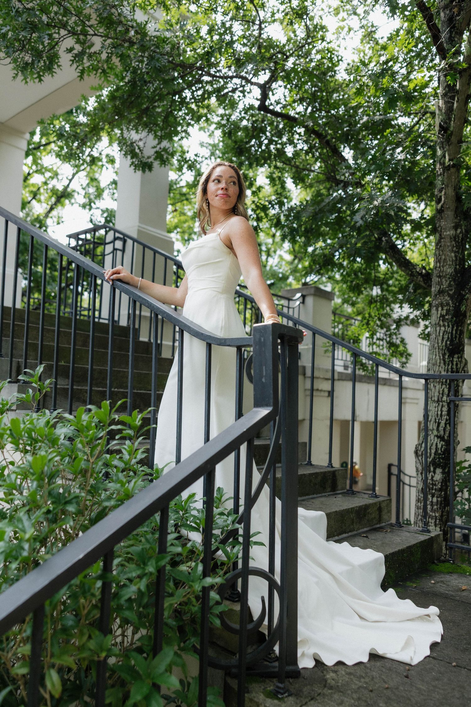 Elegant white wedding dress flowing on outdoor stone steps with black railings and green foliage.