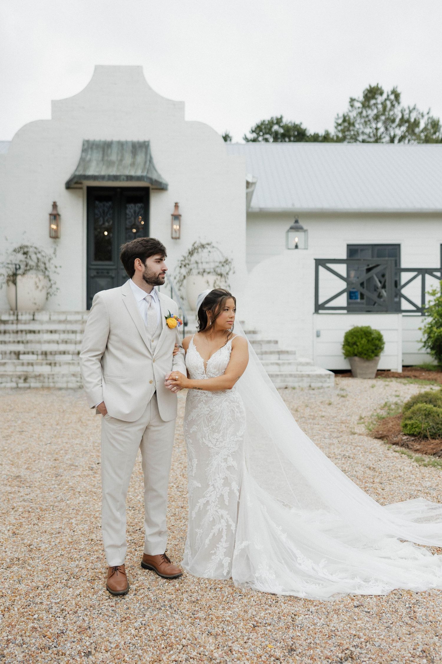 Bride and groom walking on gravel path outside white building with trailing wedding dress.