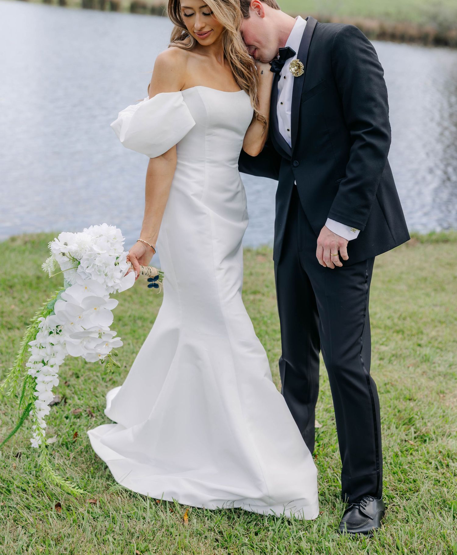 A couple in wedding attire dances together on grass near a lake.