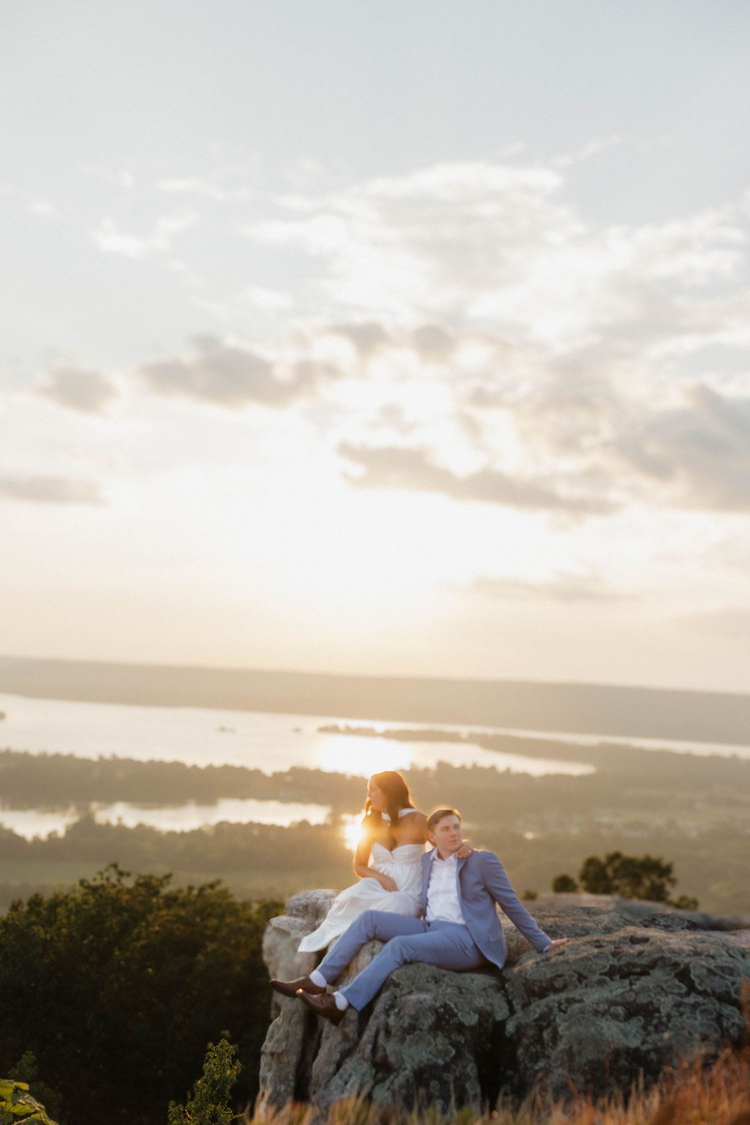 Couple relaxing on rocky outcrop during golden sunset over lake landscape.